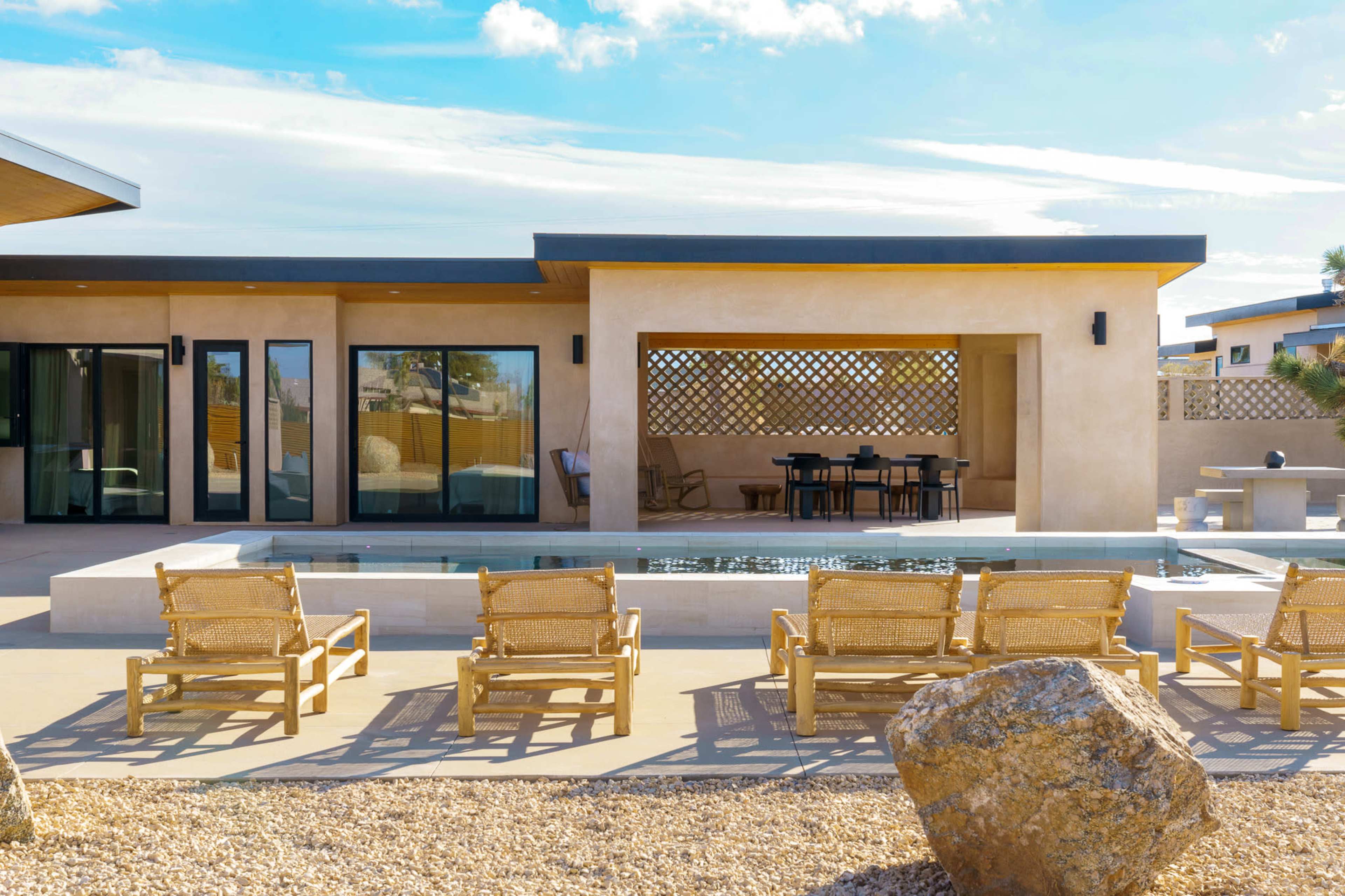 A modern, single-story house with large windows overlooks a swimming pool and a seating area surrounded by lounge chairs and a rocky landscape.