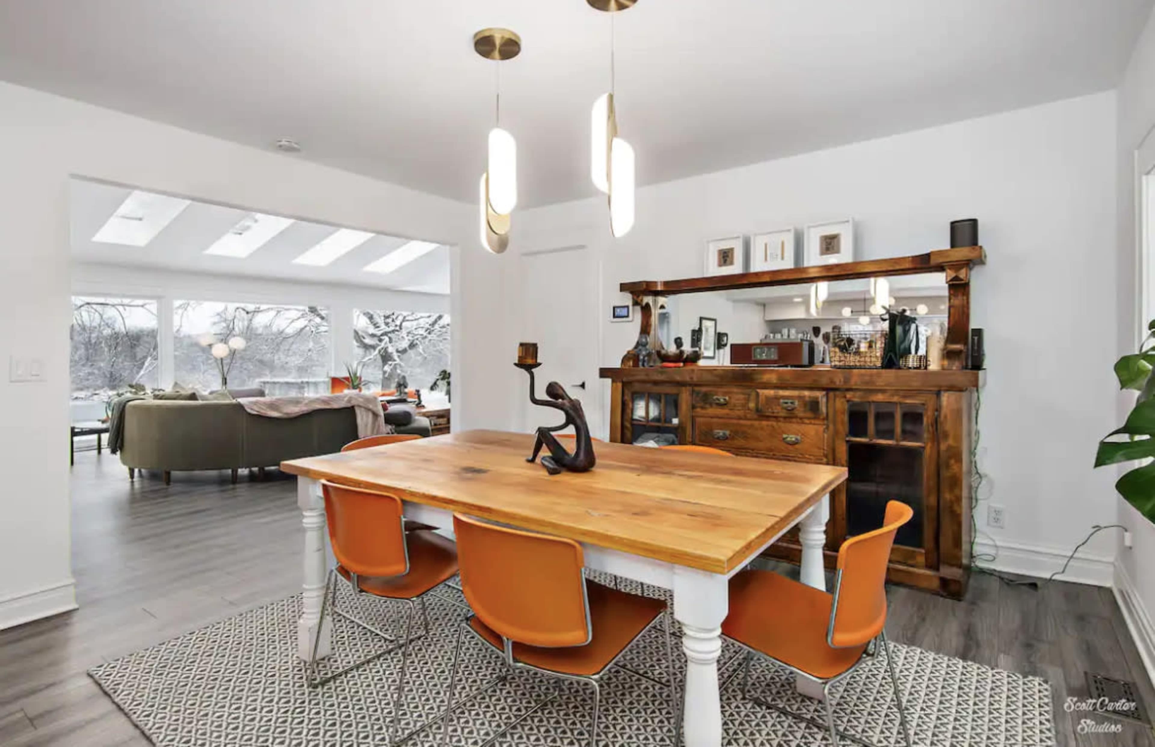 A dining area features a wooden table surrounded by orange chairs, with a sideboard and large windows overlooking a scenic view.