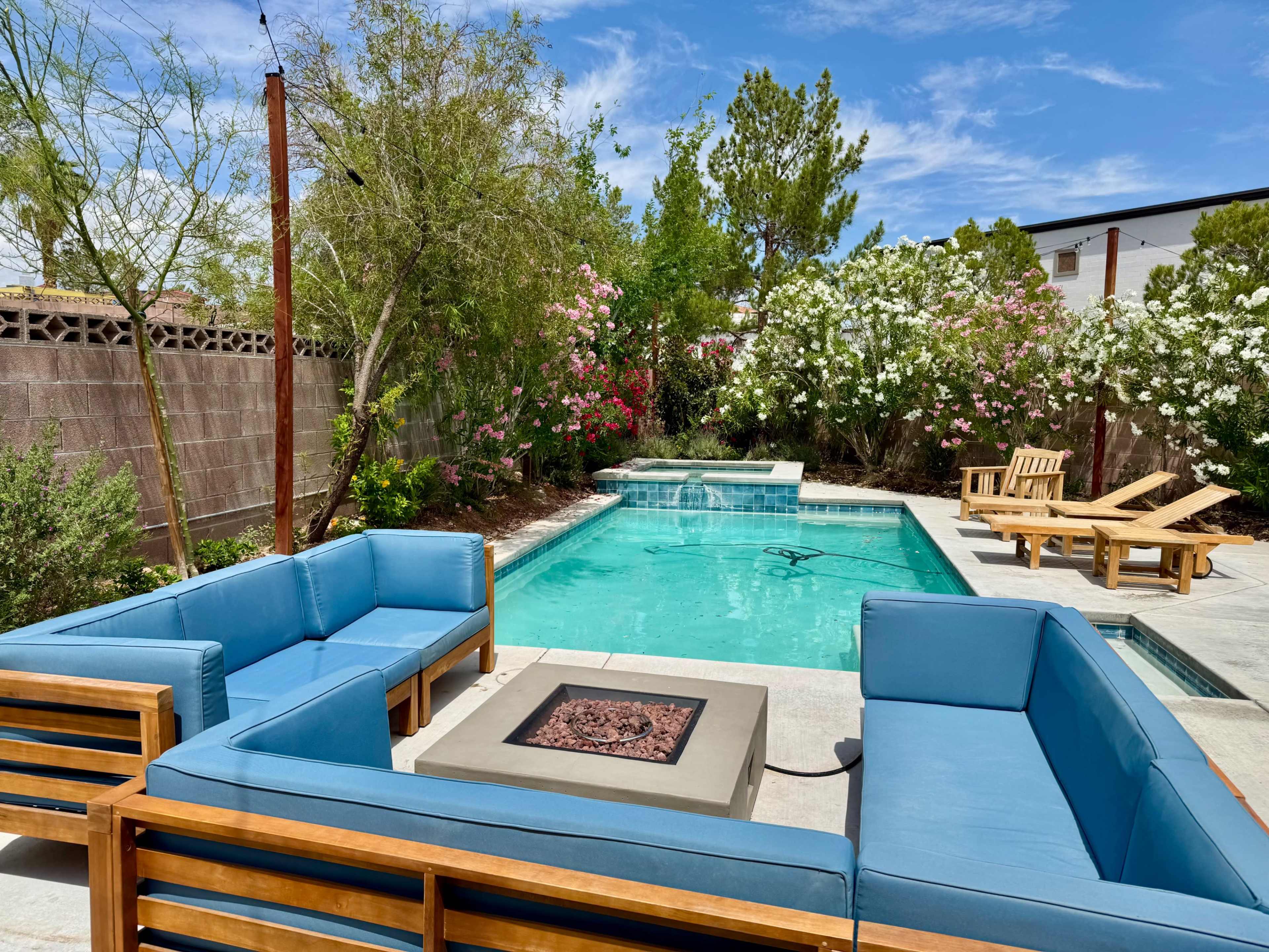 A seating area with blue cushions surrounds a rectangular swimming pool, surrounded by greenery and flowering plants.