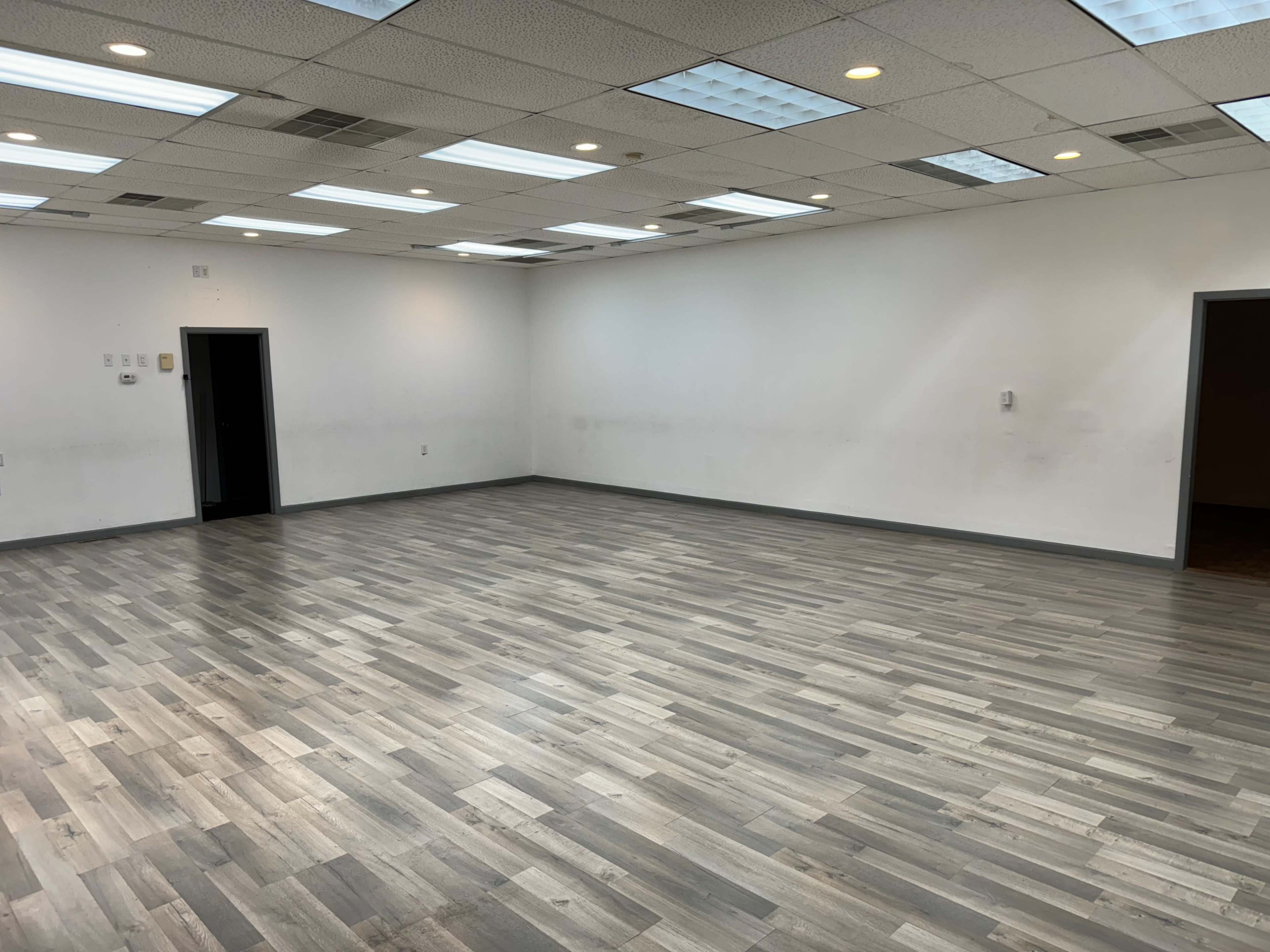 A spacious, empty room with gray laminate flooring and white walls, featuring several ceiling lights and two doorways.