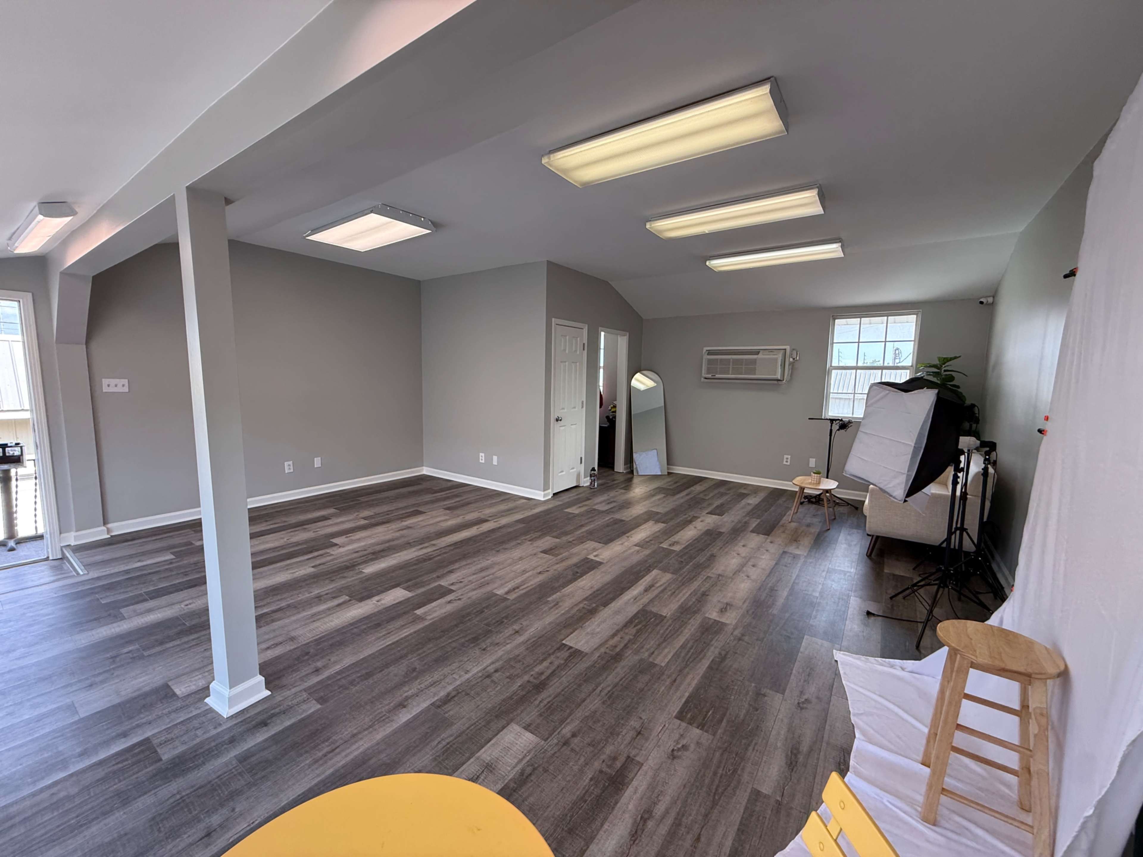 The image shows a spacious, empty room with gray walls, wood-patterned flooring, and several ceiling lights, featuring a door and a window with an air conditioning unit.