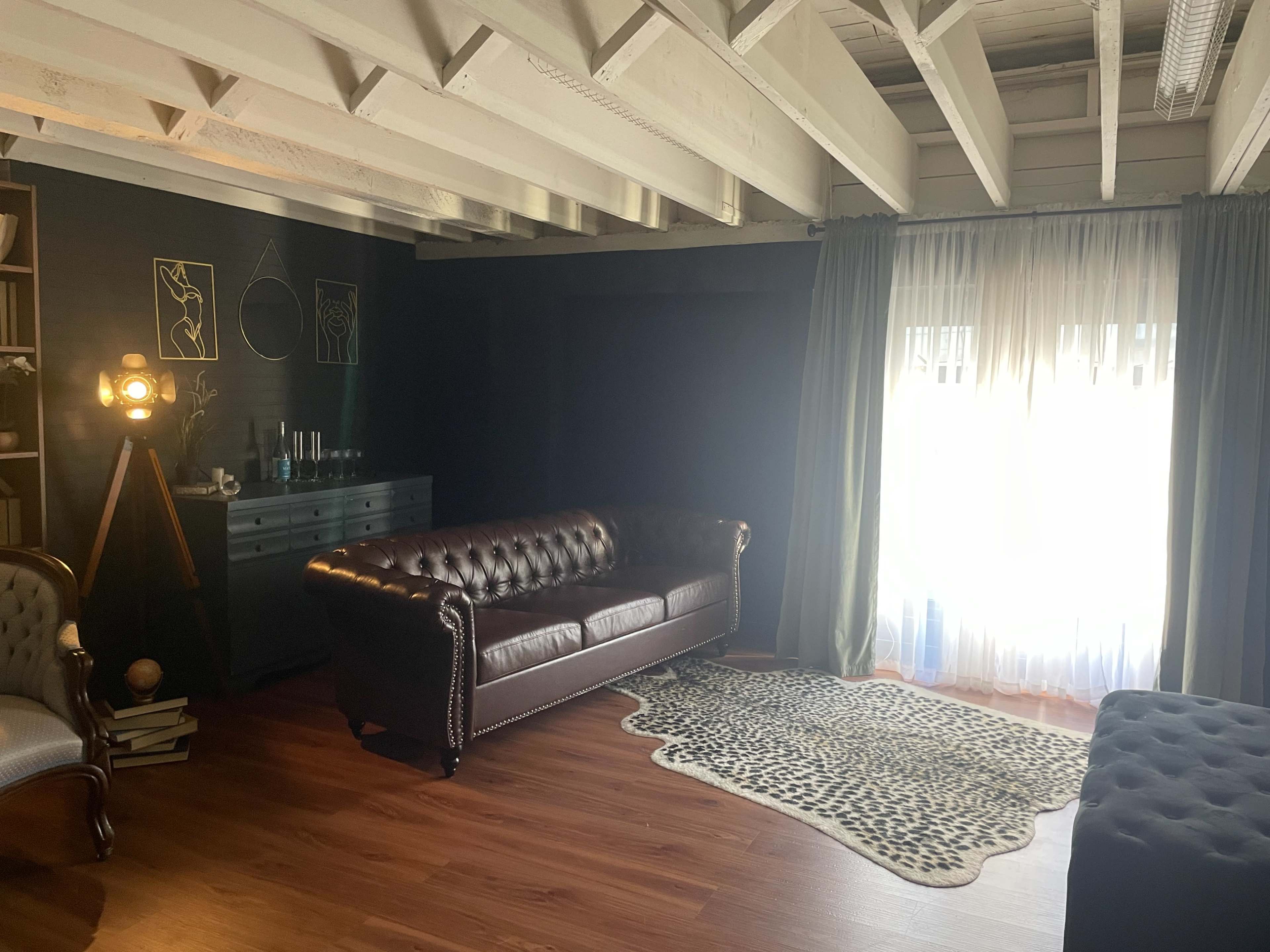 The image shows a furnished living room featuring a brown leather chesterfield sofa, a patterned rug, and large windows with sheer curtains.