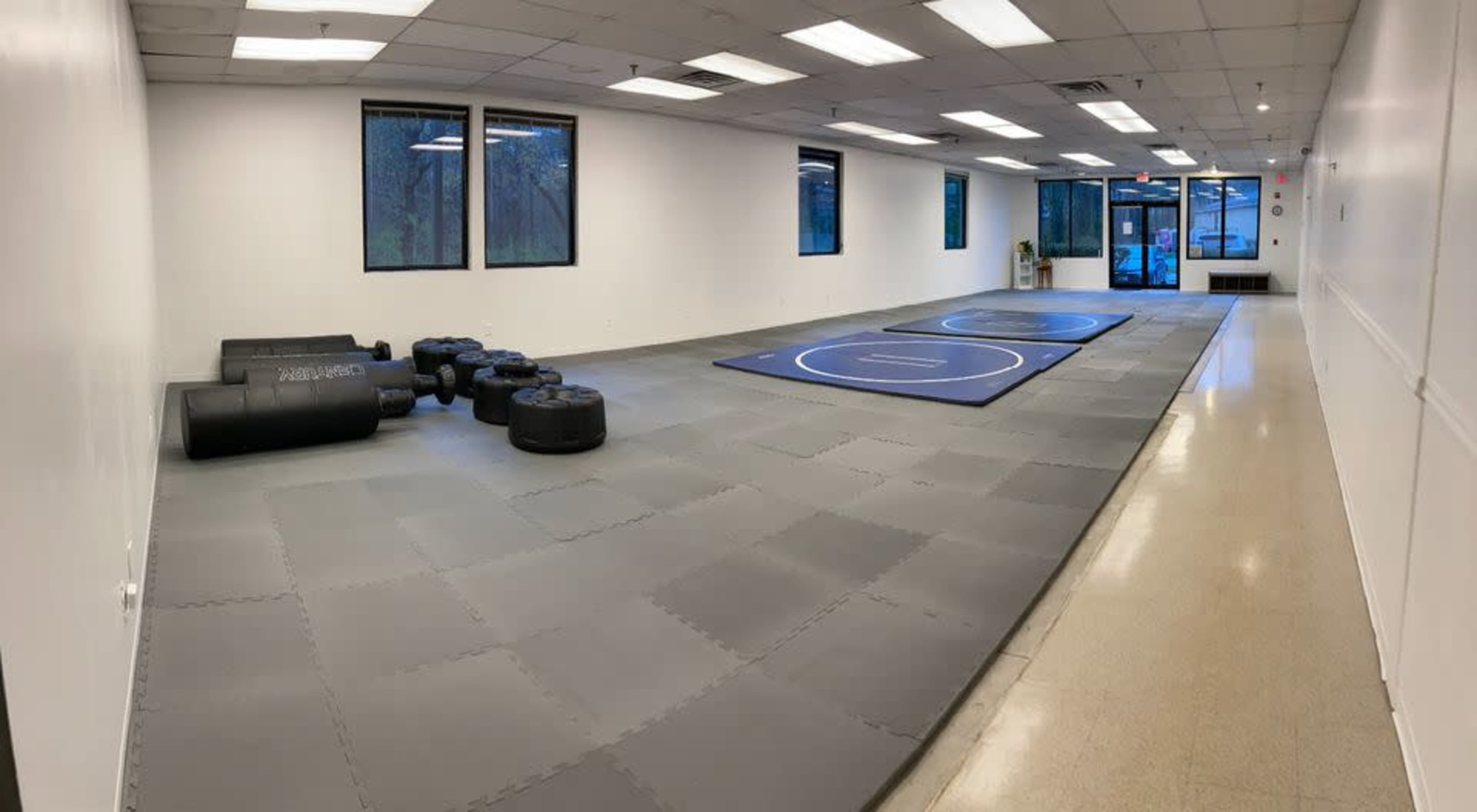 A spacious, empty training room with gray mats, two circular wrestling rings, and black practice equipment arranged on one side.