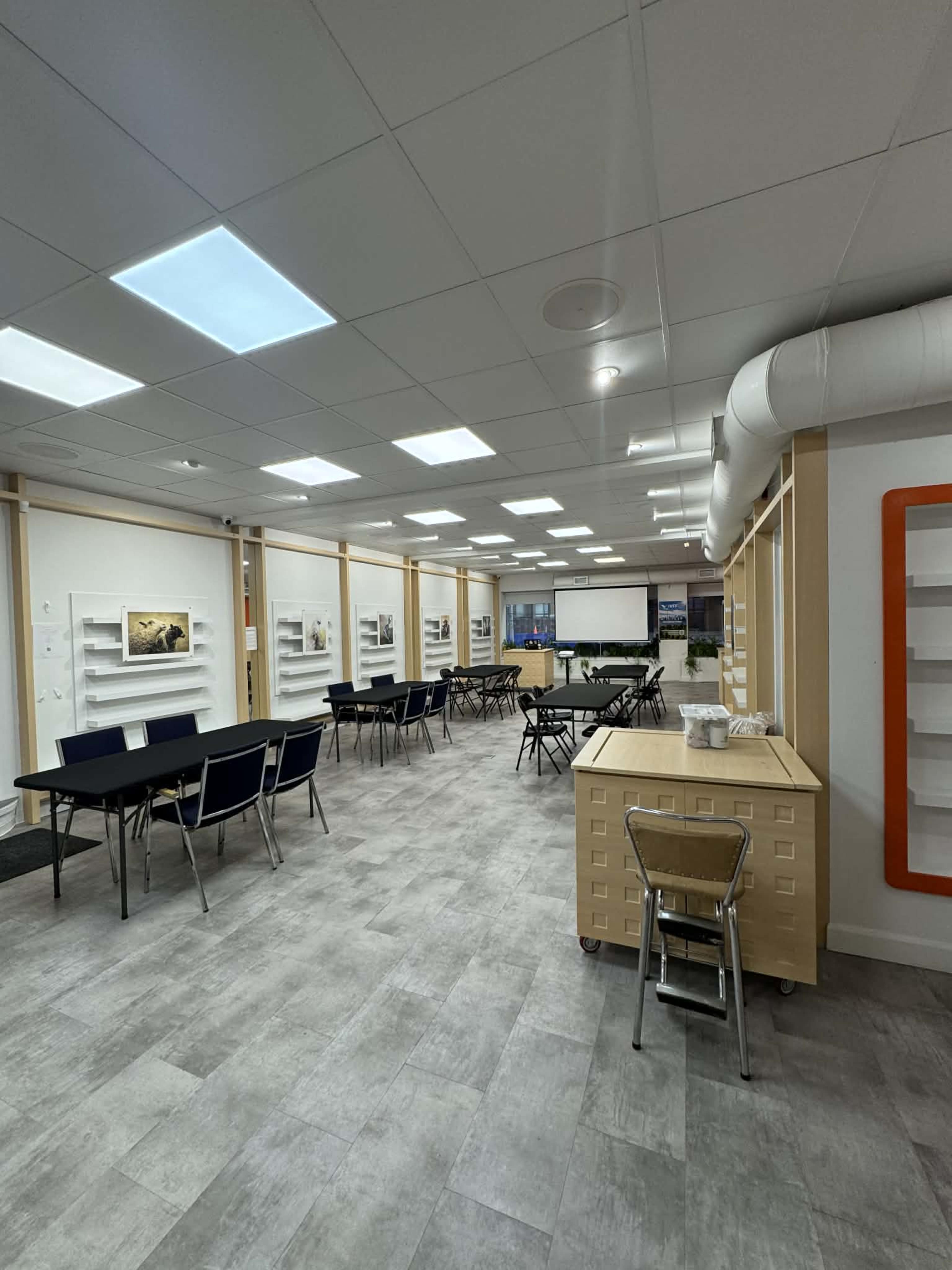 The image shows a spacious room set up for a meeting or event, featuring rows of tables and chairs, a large screen at the front, and walls adorned with framed images.