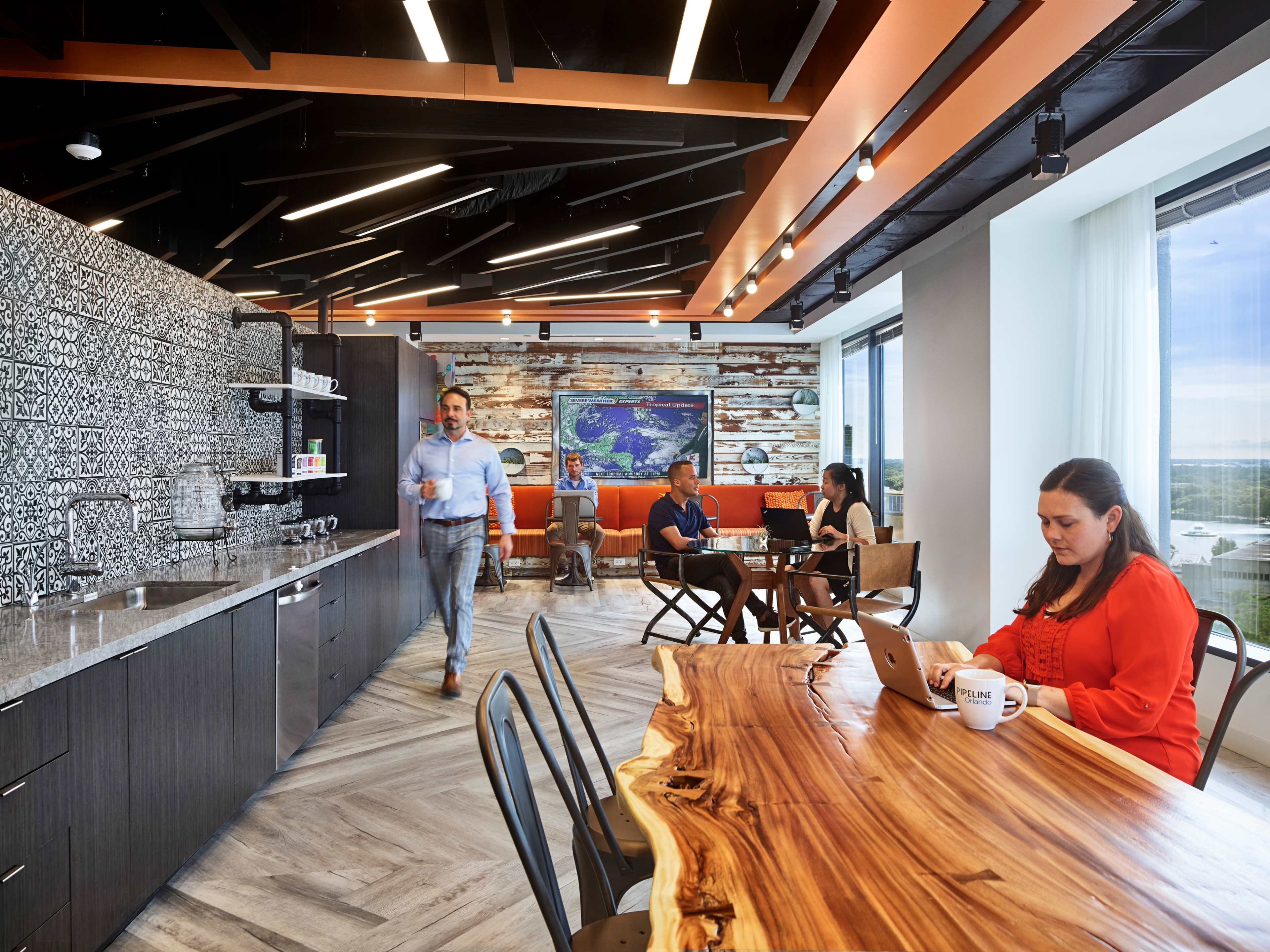 A modern office break room with a kitchen area, a large wooden table, seating areas, and people engaged in conversation and work.