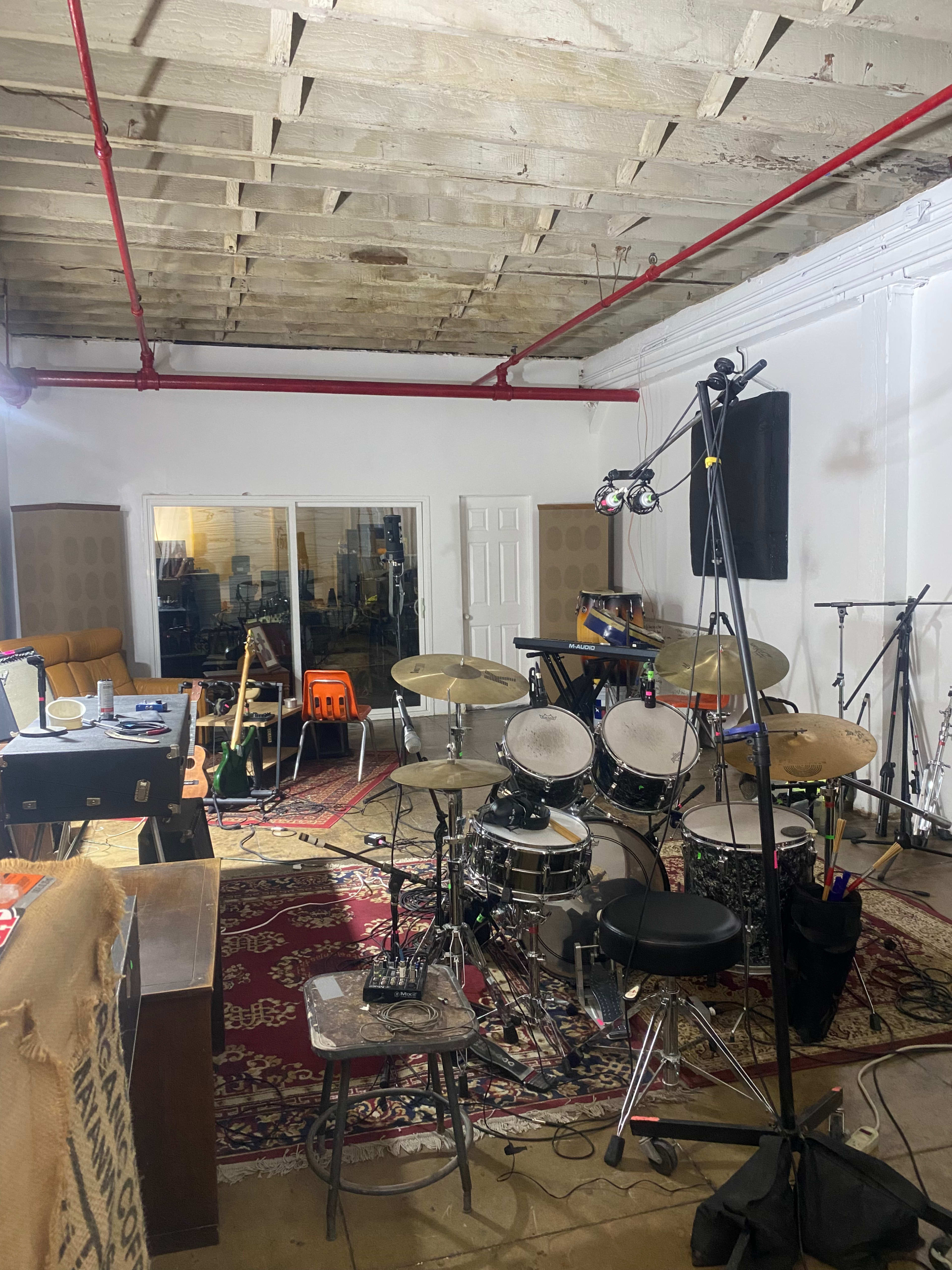 The image shows a music rehearsal space featuring a drum set, microphones, and various instruments arranged around a carpeted area with white walls and exposed ceilings.