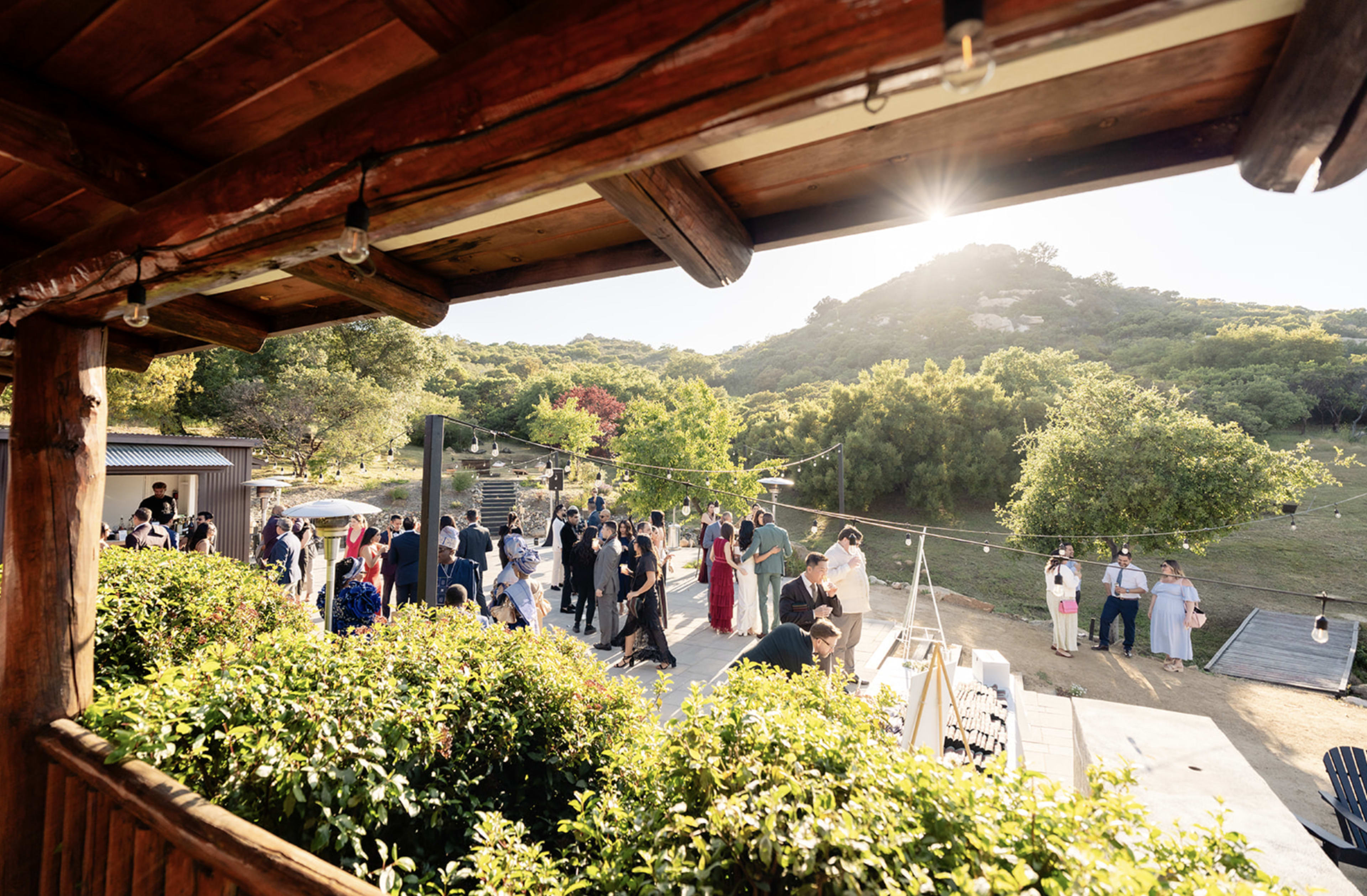 A gathering of people is taking place outdoors, surrounded by greenery and wooden structures, with sunlight streaming in from the upper left.