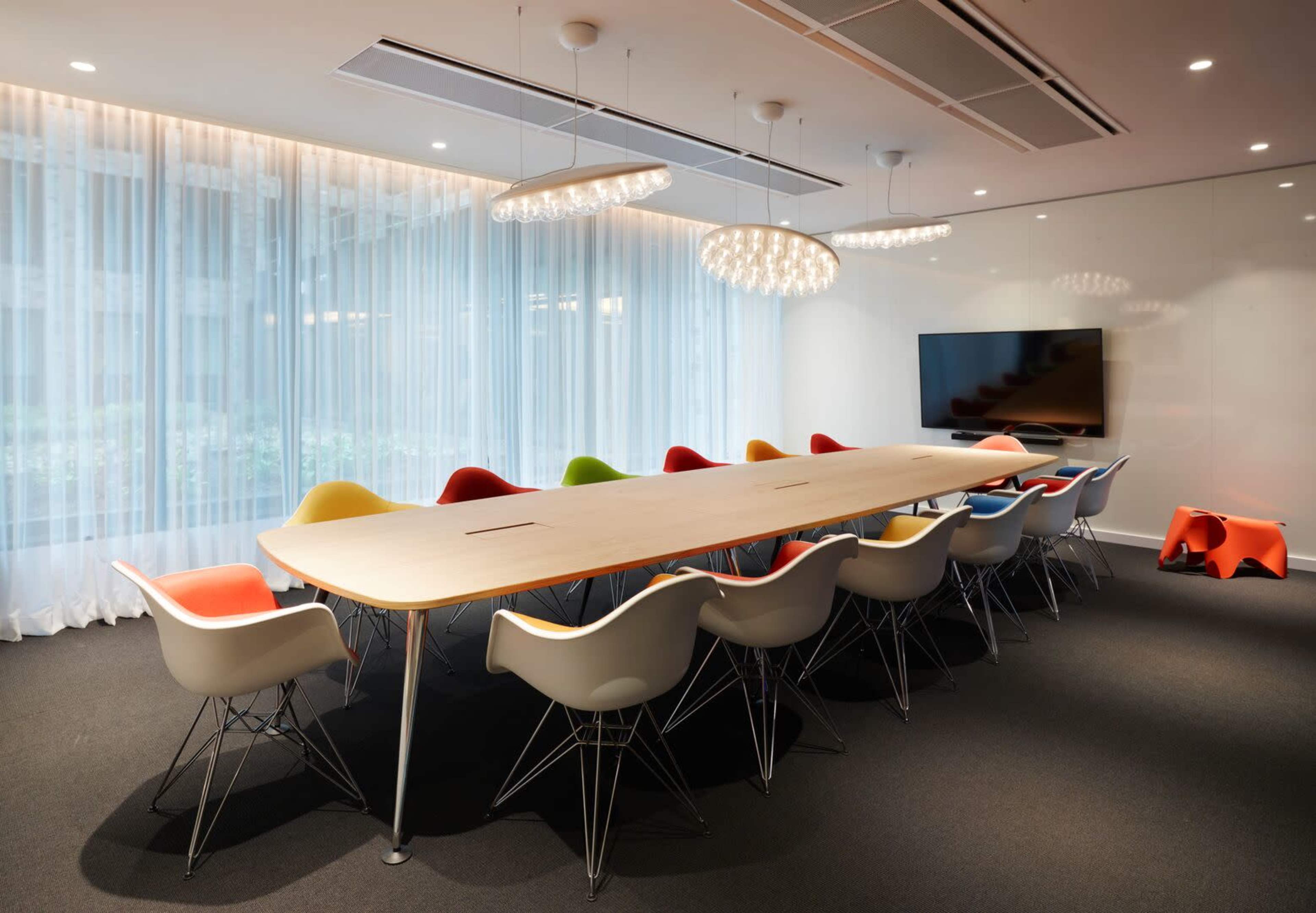 A large conference room features a long wooden table surrounded by colorful Eames-style chairs and a wall-mounted television.