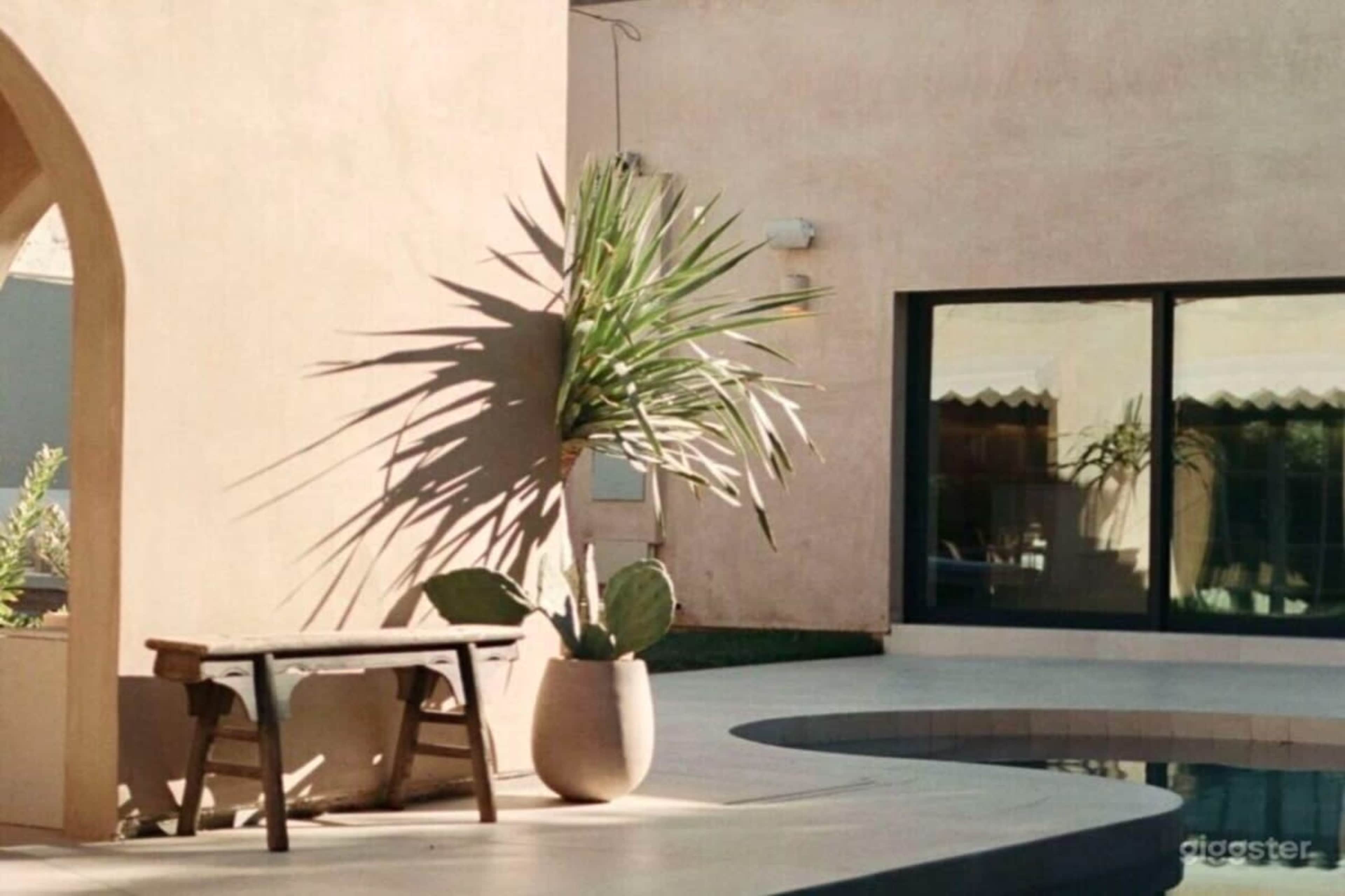 The scene features a minimalist outdoor space with a small wooden table beside a large potted plant and a swimming pool, set against a smooth, beige wall.
