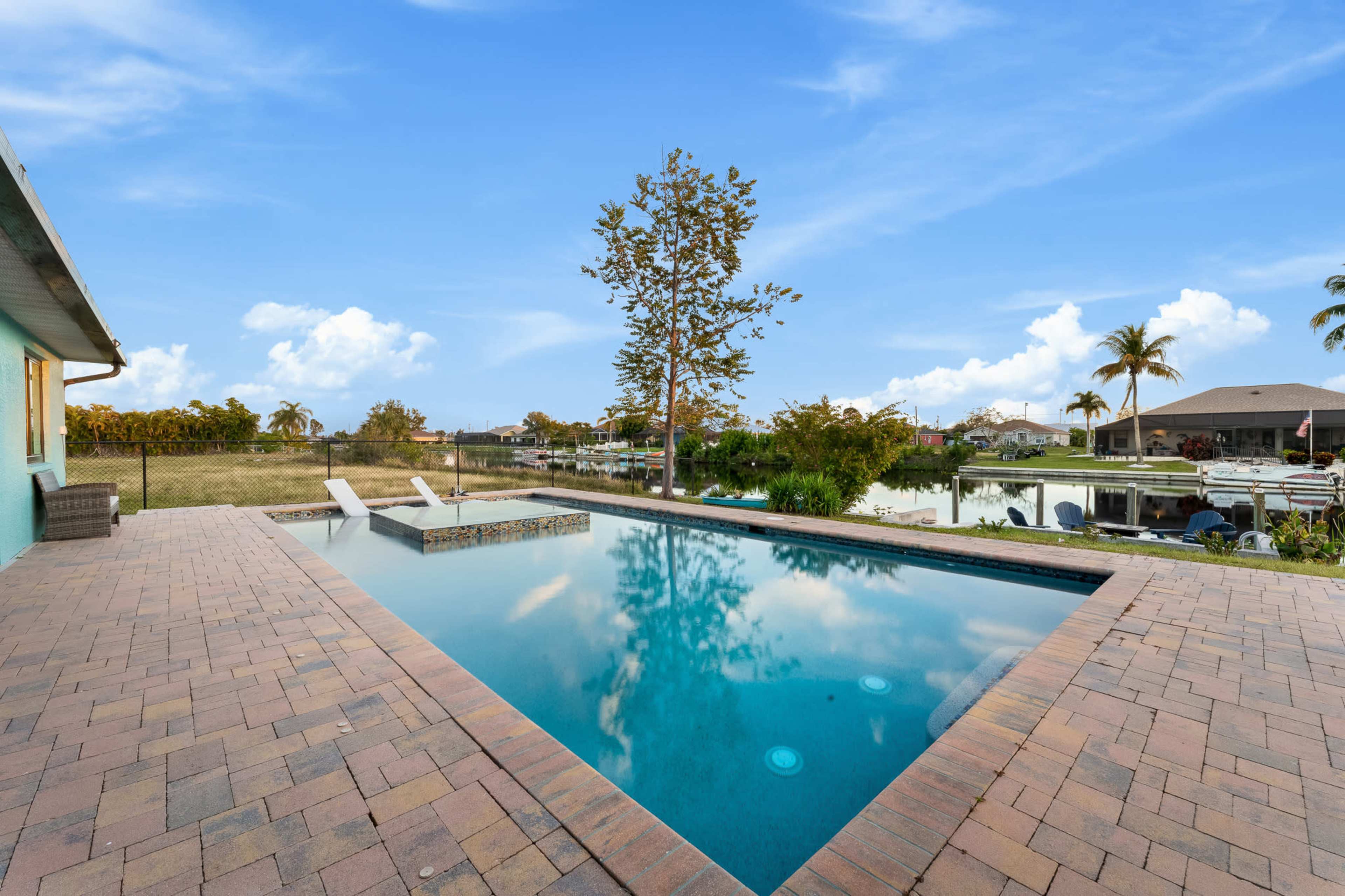A rectangular swimming pool with two lounge chairs is surrounded by landscaped greenery and houses in the background under a clear blue sky.