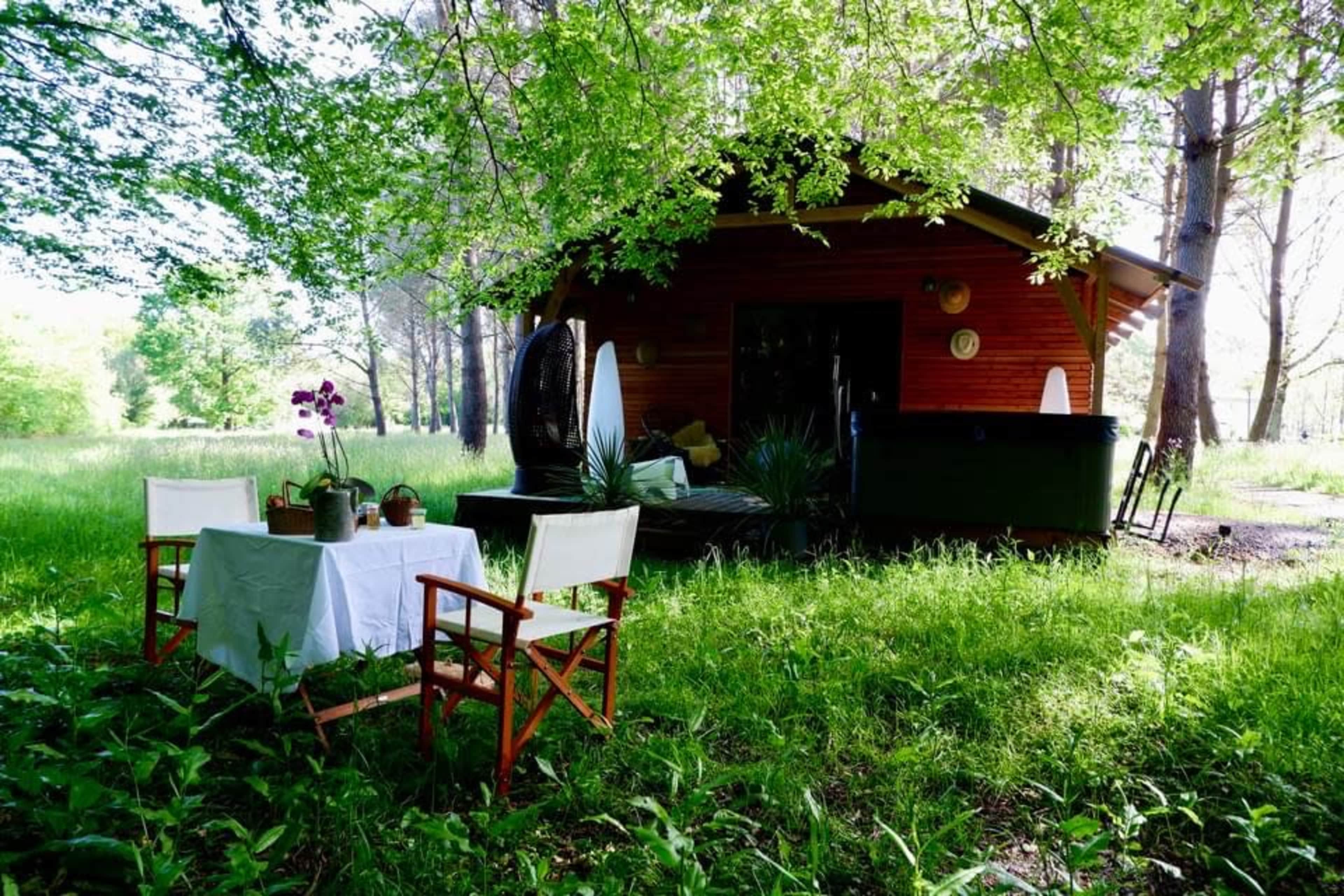 A small wooden cabin sits among tall trees, with a table and chairs set up on the grassy area nearby.