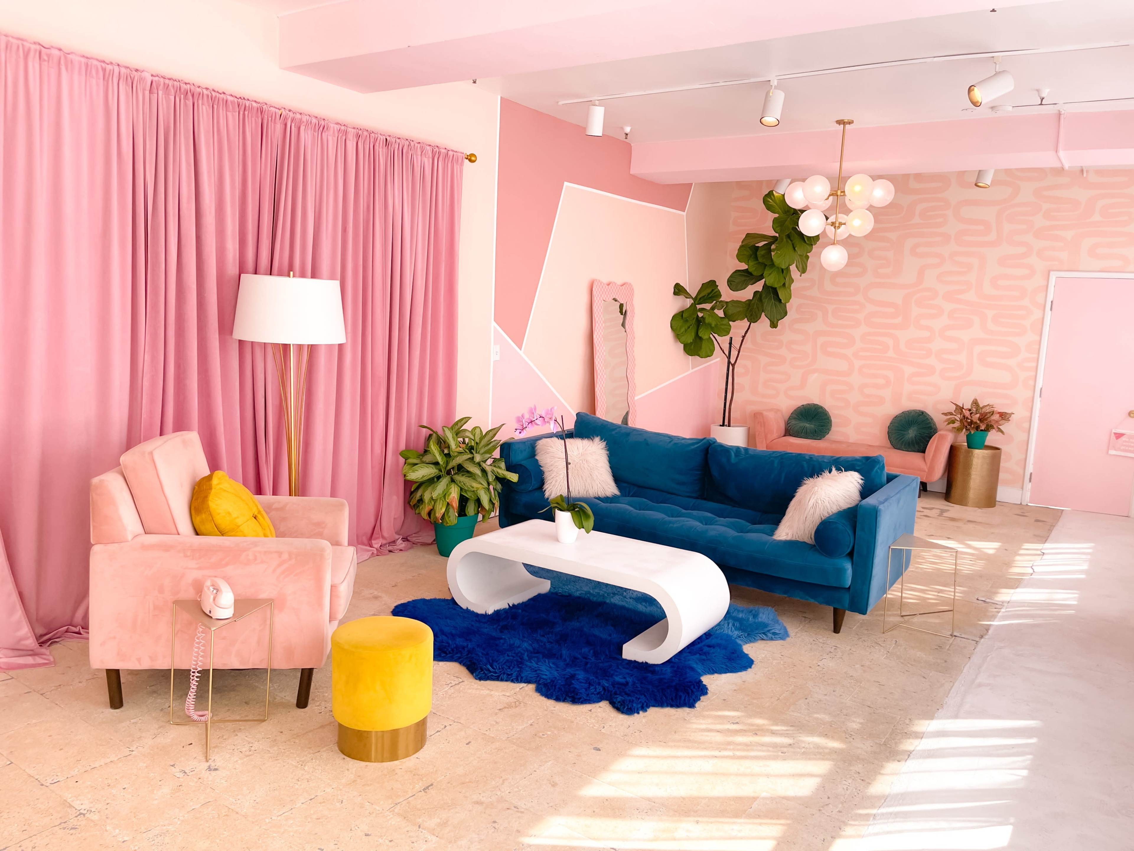 A colorful living room features a blue sofa, pink armchair, decorative plants, and a white coffee table against a backdrop of pink curtains and geometric wall patterns.