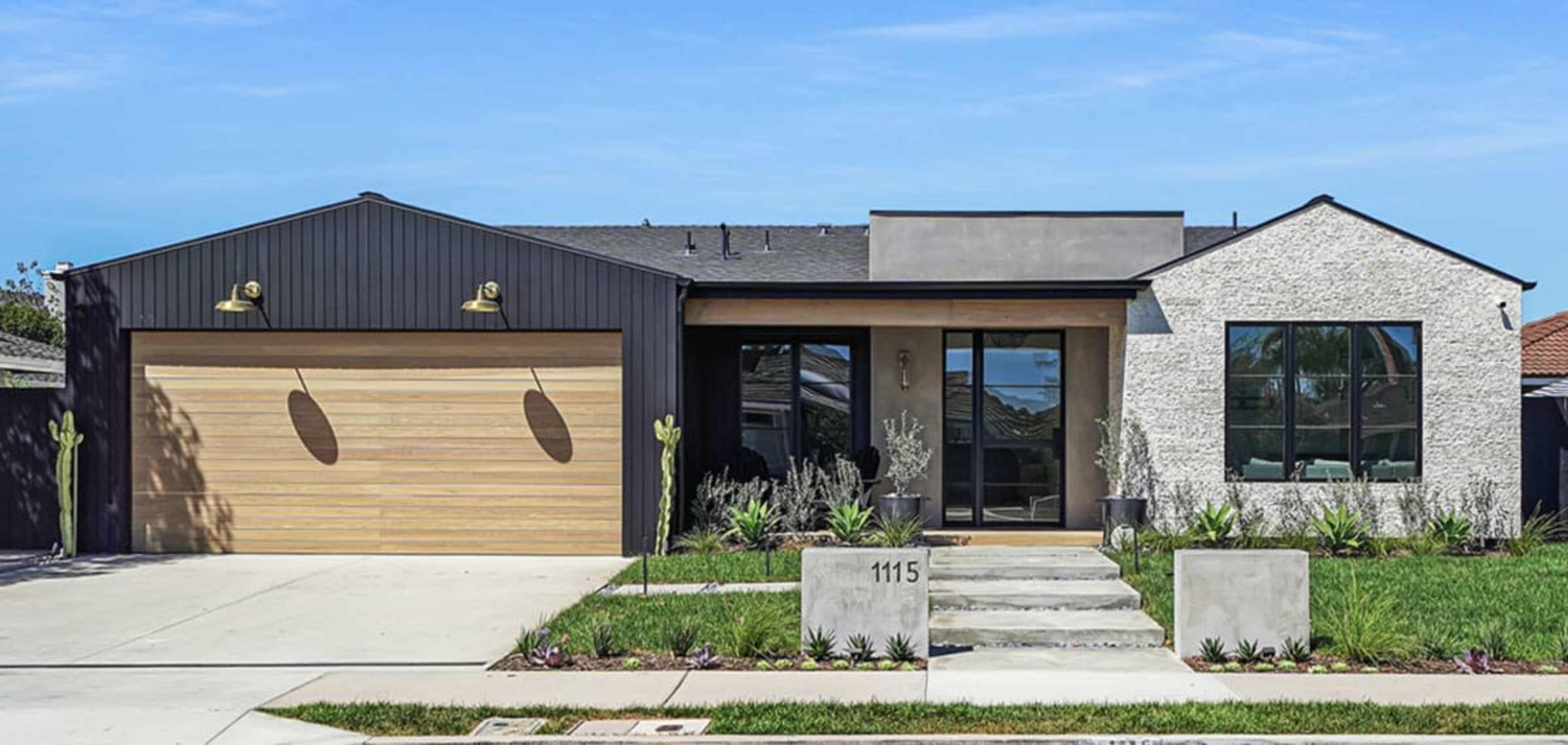 The image depicts a modern single-story house with a mix of textured exterior materials, large windows, and a wooden garage door.