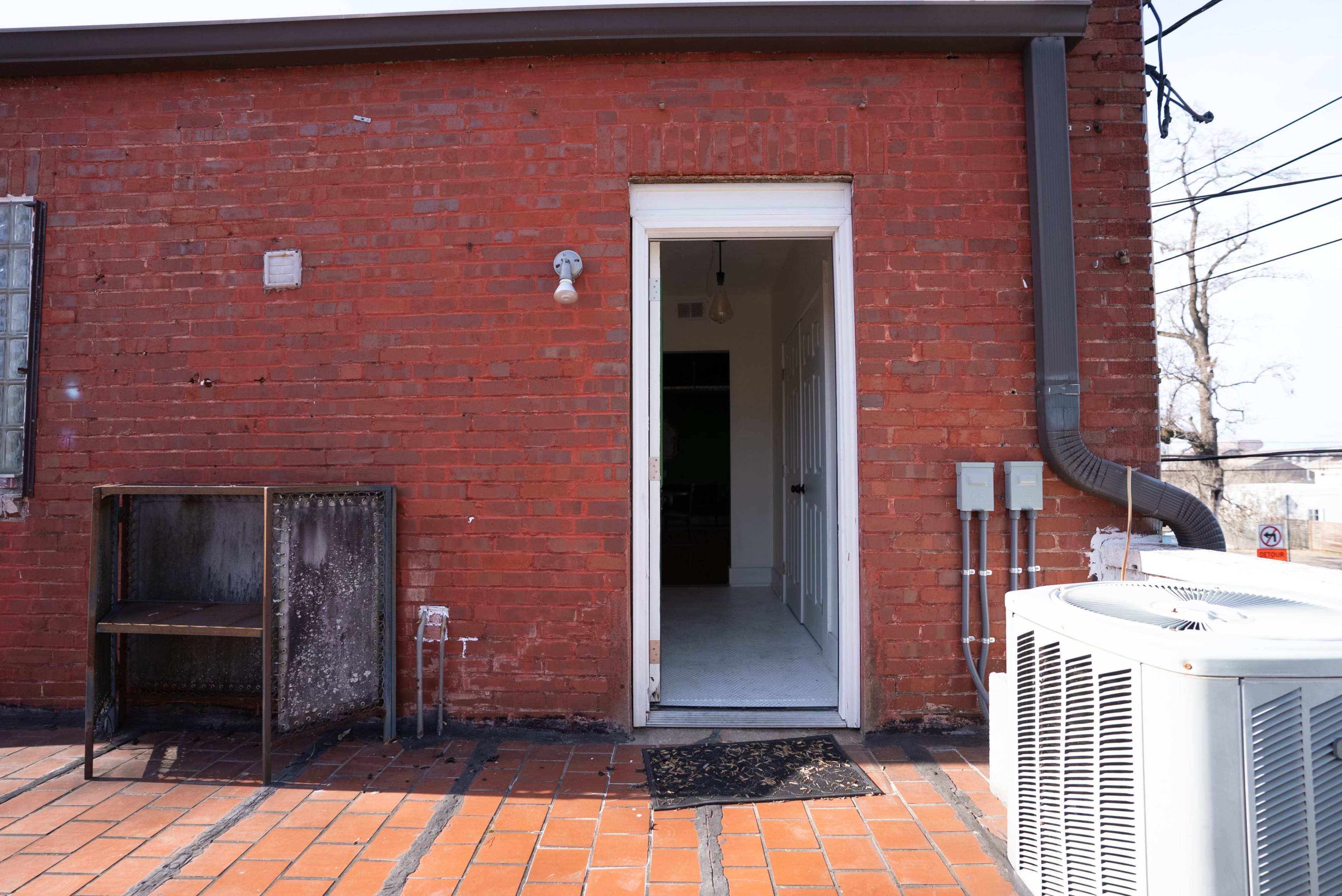 The image shows a brick exterior wall with an open door leading into a white interior space, accompanied by a metal table, an air conditioning unit, and visible power lines.