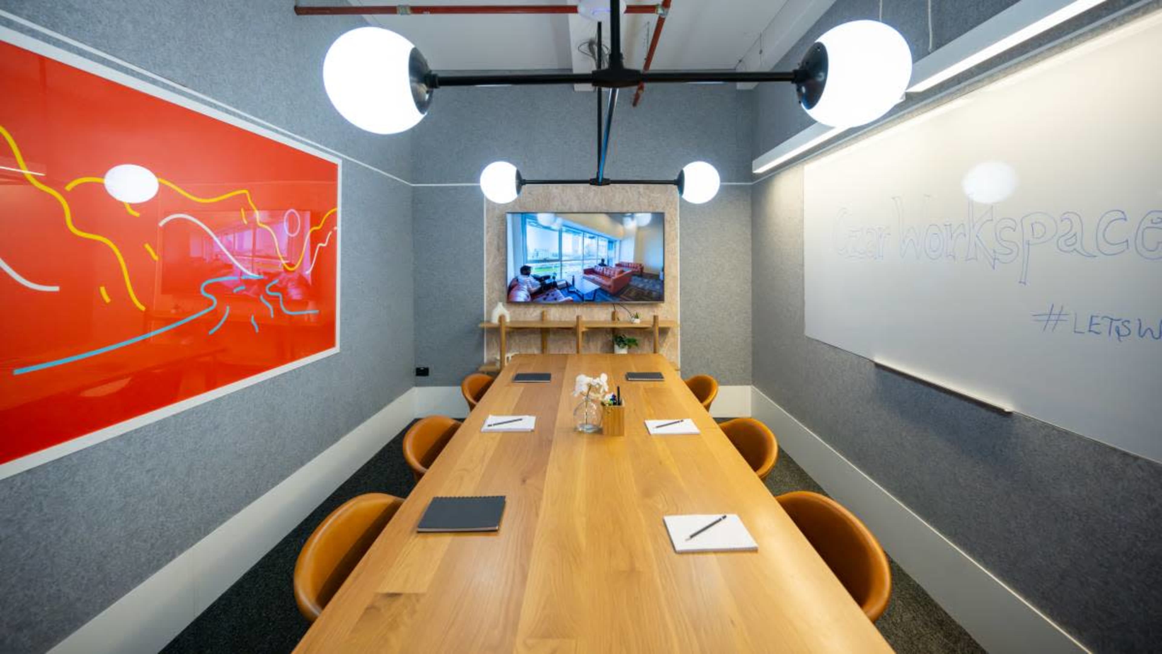 A modern conference room features a long wooden table surrounded by orange chairs, with a television and whiteboard on the walls, and a vibrant artwork displayed.