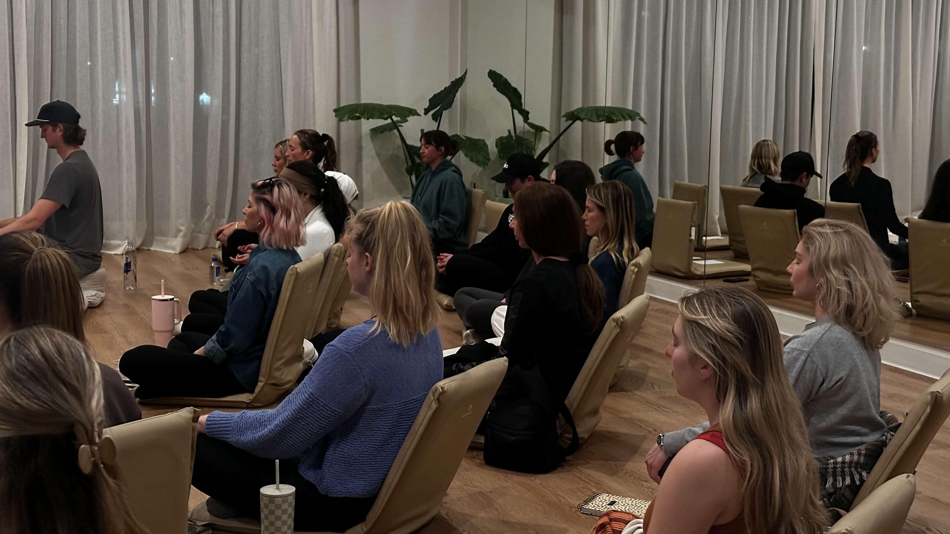 A group of people sits in a neat arrangement of chairs in a room with large curtains and potted plants.