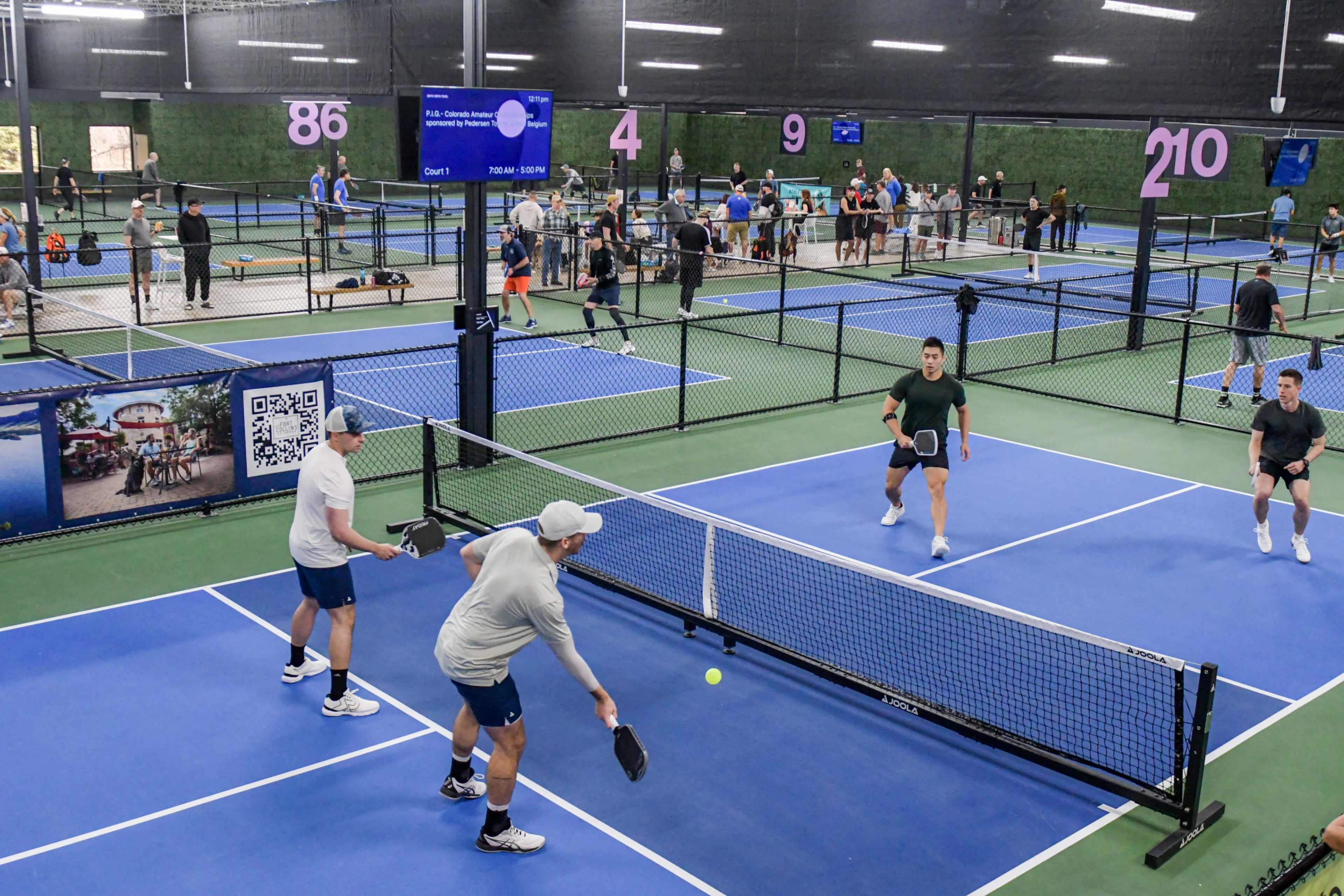 Players are engaged in multiple games of pickleball in a sports facility with several courts and spectators.