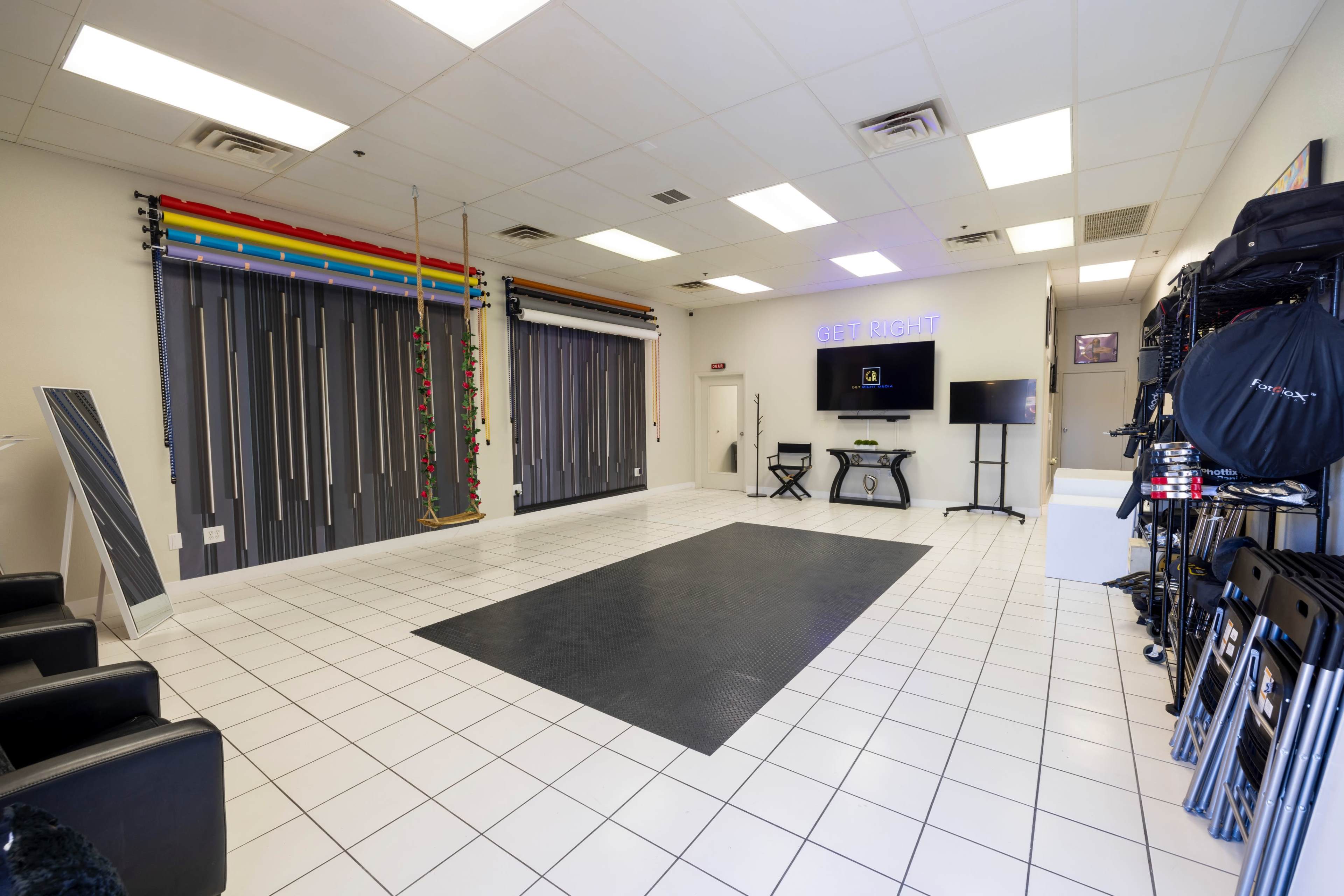 The image shows a spacious, well-lit studio with a black mat on the floor, colorful backdrop strips on the wall, and various equipment neatly arranged on the side.