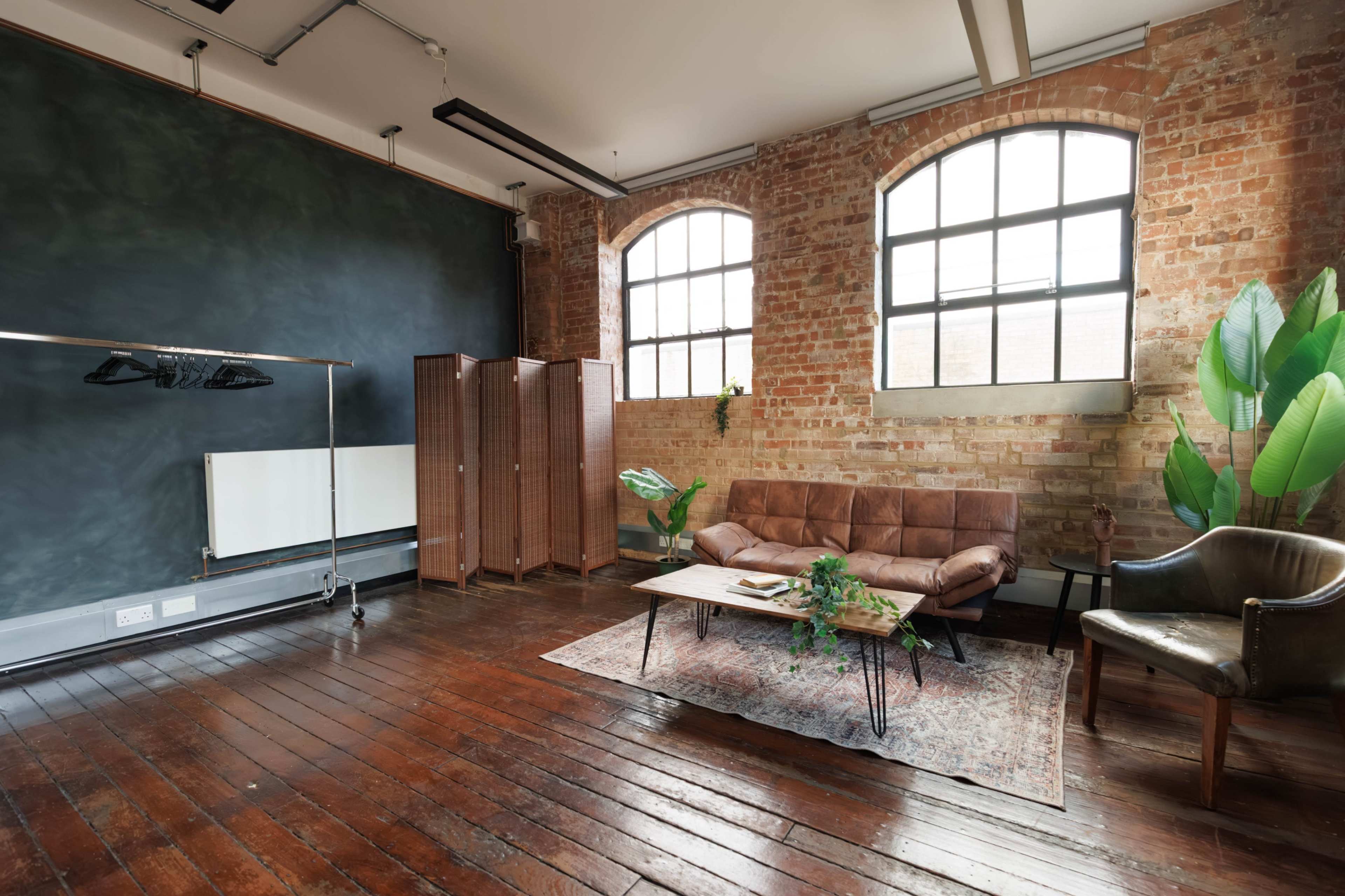 The image shows a spacious room with exposed brick walls, large windows, a brown leather sofa, and a wooden coffee table, accompanied by potted plants and a clothing rack.