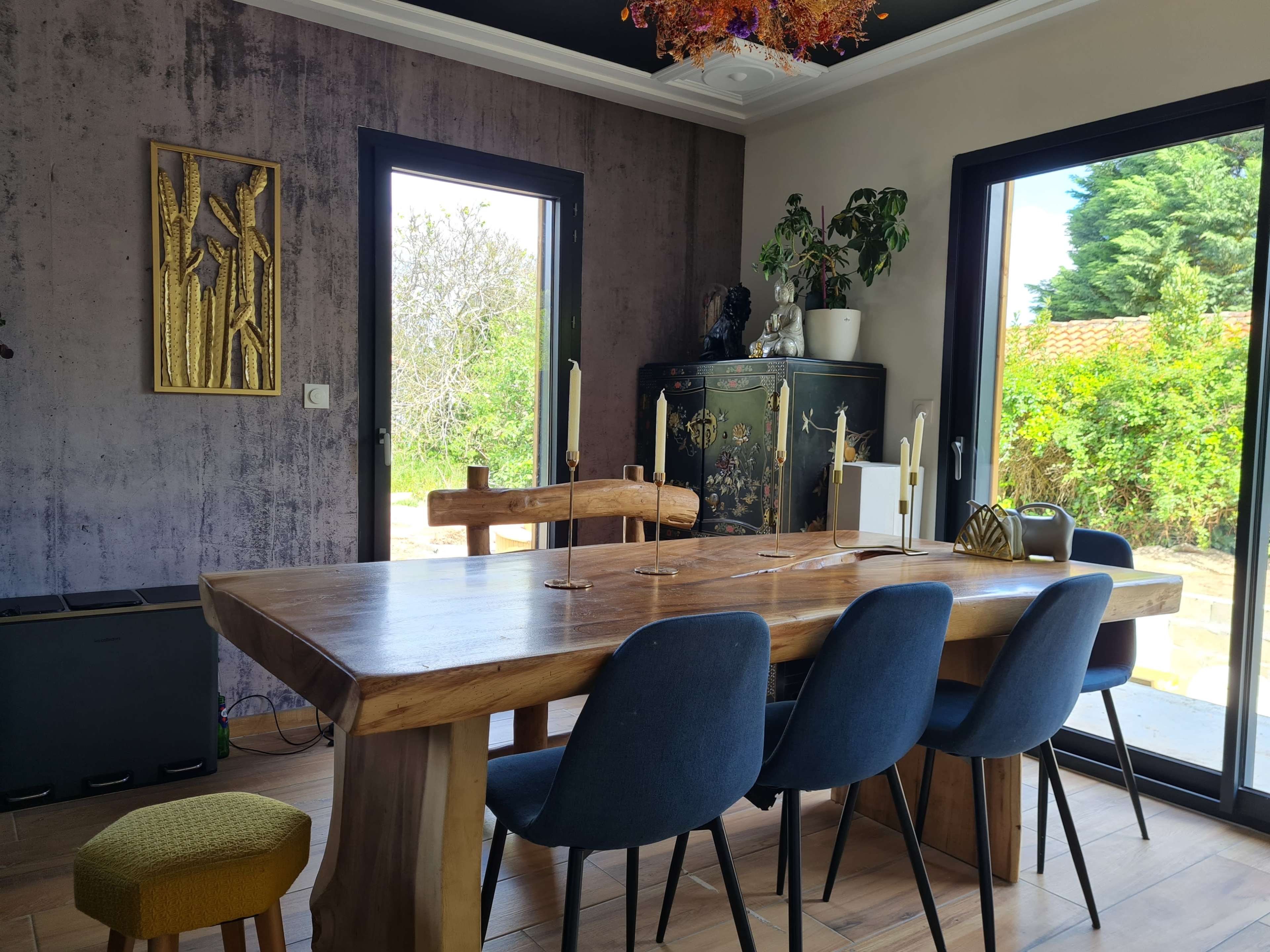 A wooden dining table with candles sits in a room featuring large windows and a mix of decor elements, including a piece of wall art and a plant.