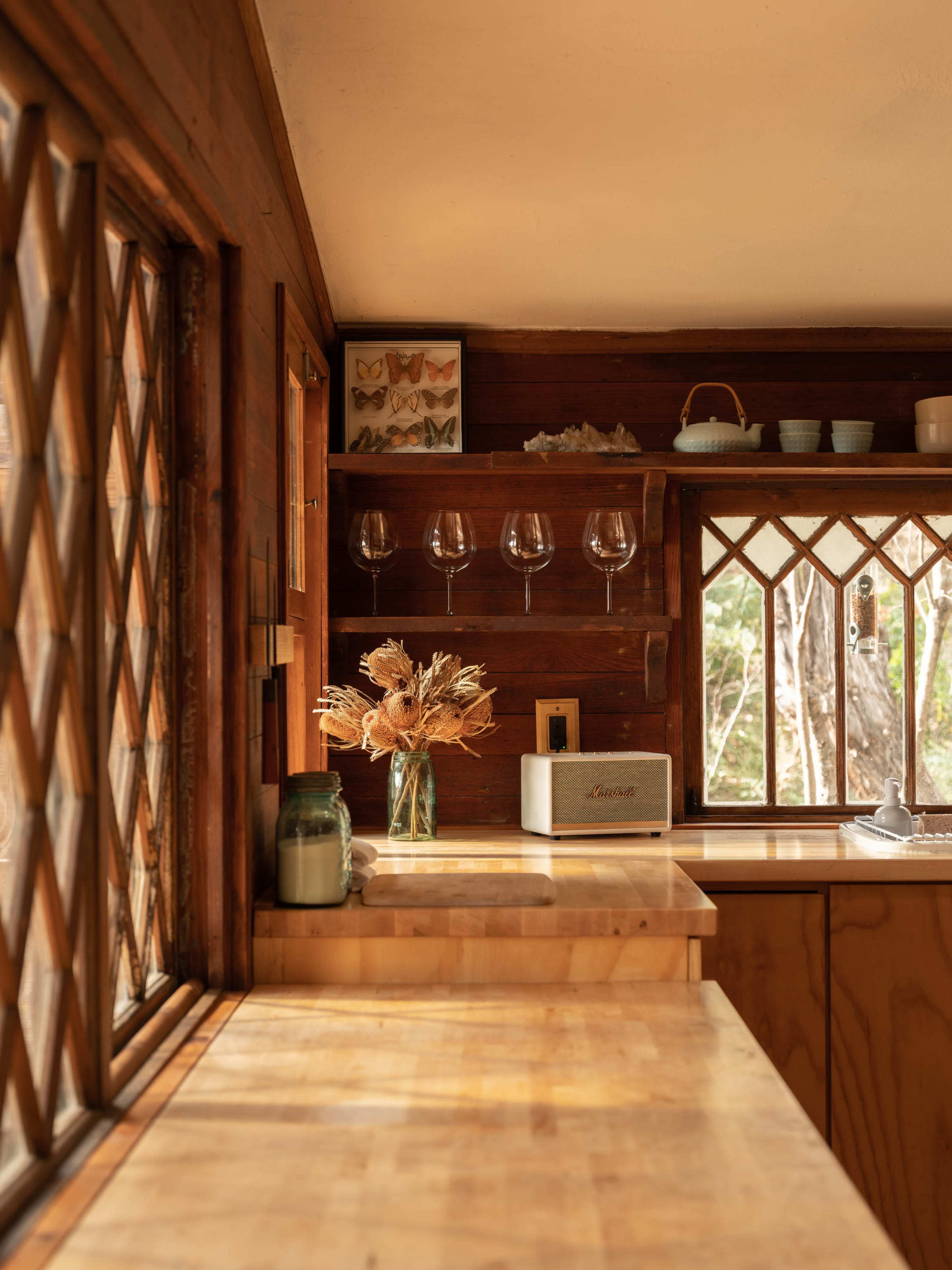 A light-filled kitchen features wooden countertops, a vase of dried flowers, and glassware displayed on a shelf.