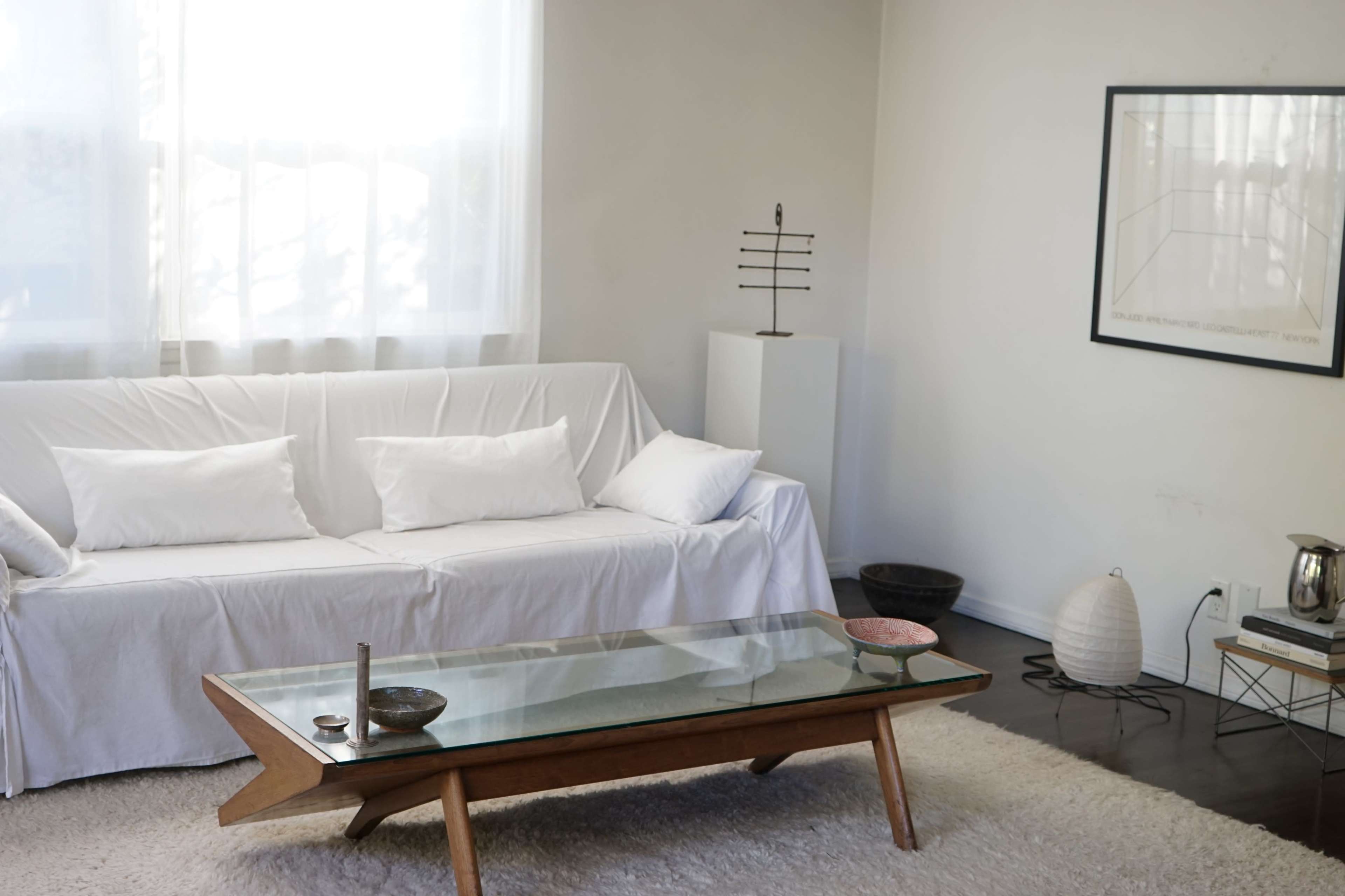 A minimalist living room featuring a white sofa, a glass coffee table with wooden legs, and a small decorative item on the table.