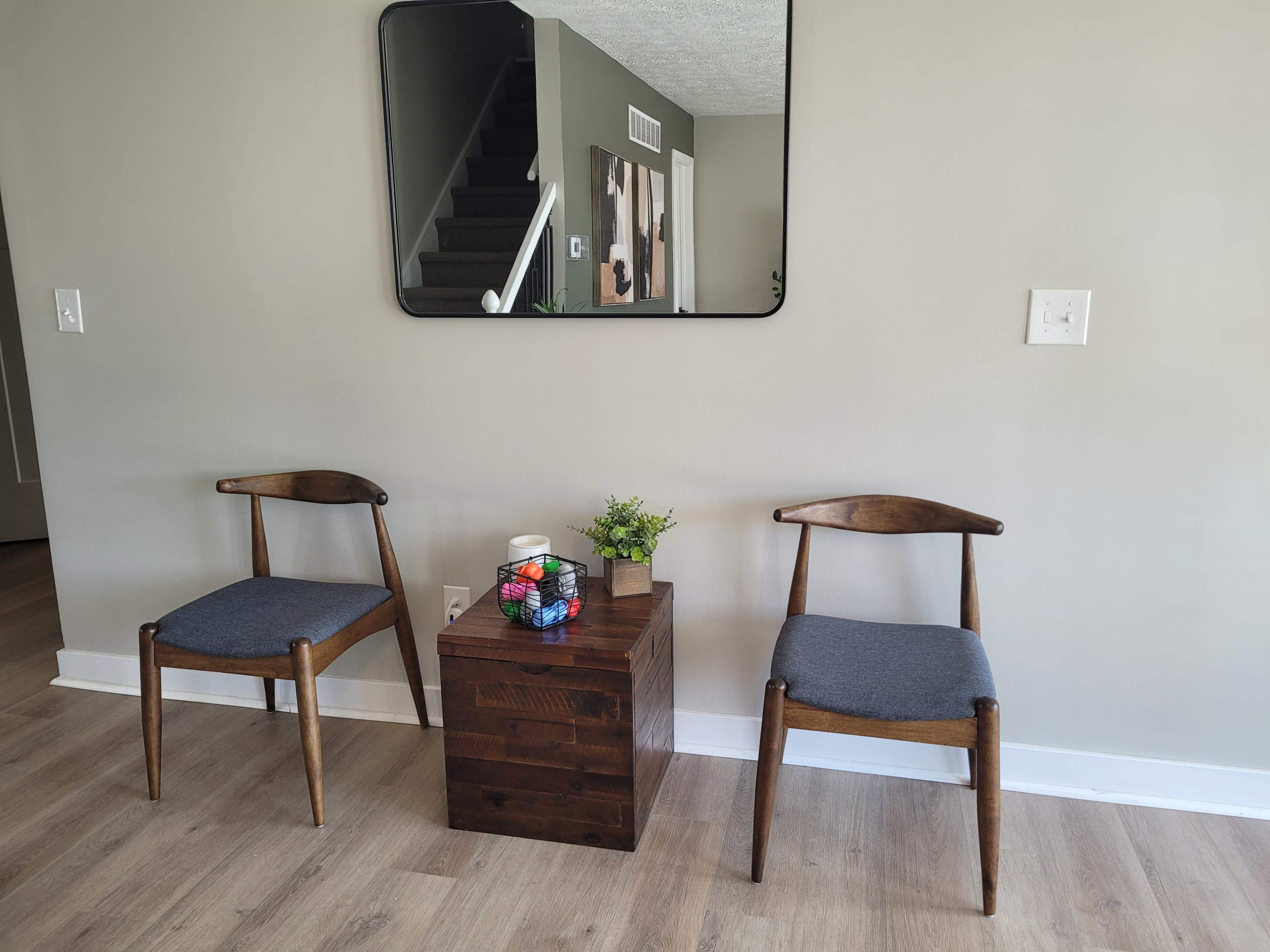 The image shows a small seating area with two wooden chairs and a square wooden side table, featuring a plant and a colorful Rubik's Cube, against a light gray wall with a mirror above.