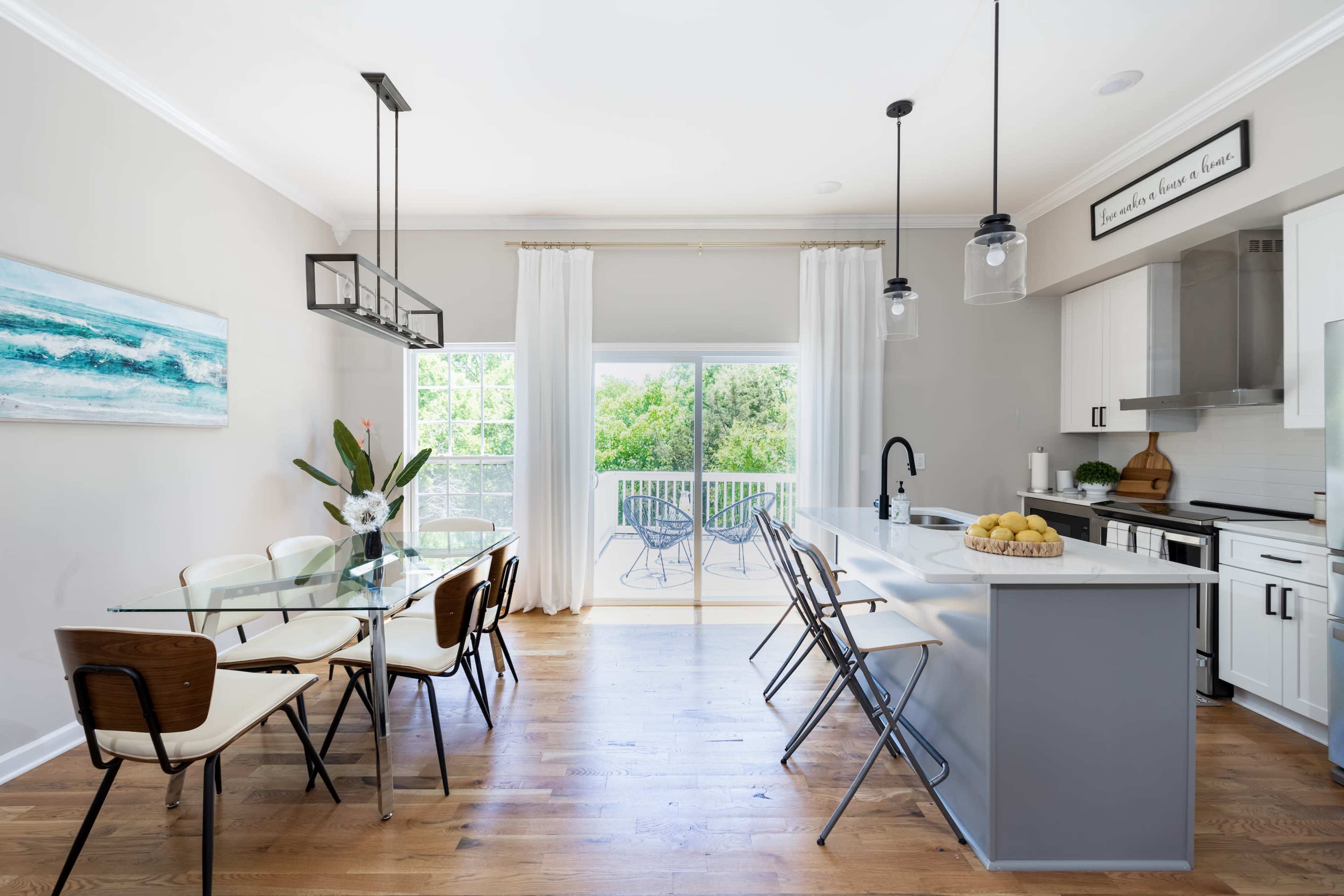 A modern kitchen-dining area features a glass dining table, wooden chairs, and a sliding door that opens to a balcony overlooking greenery.