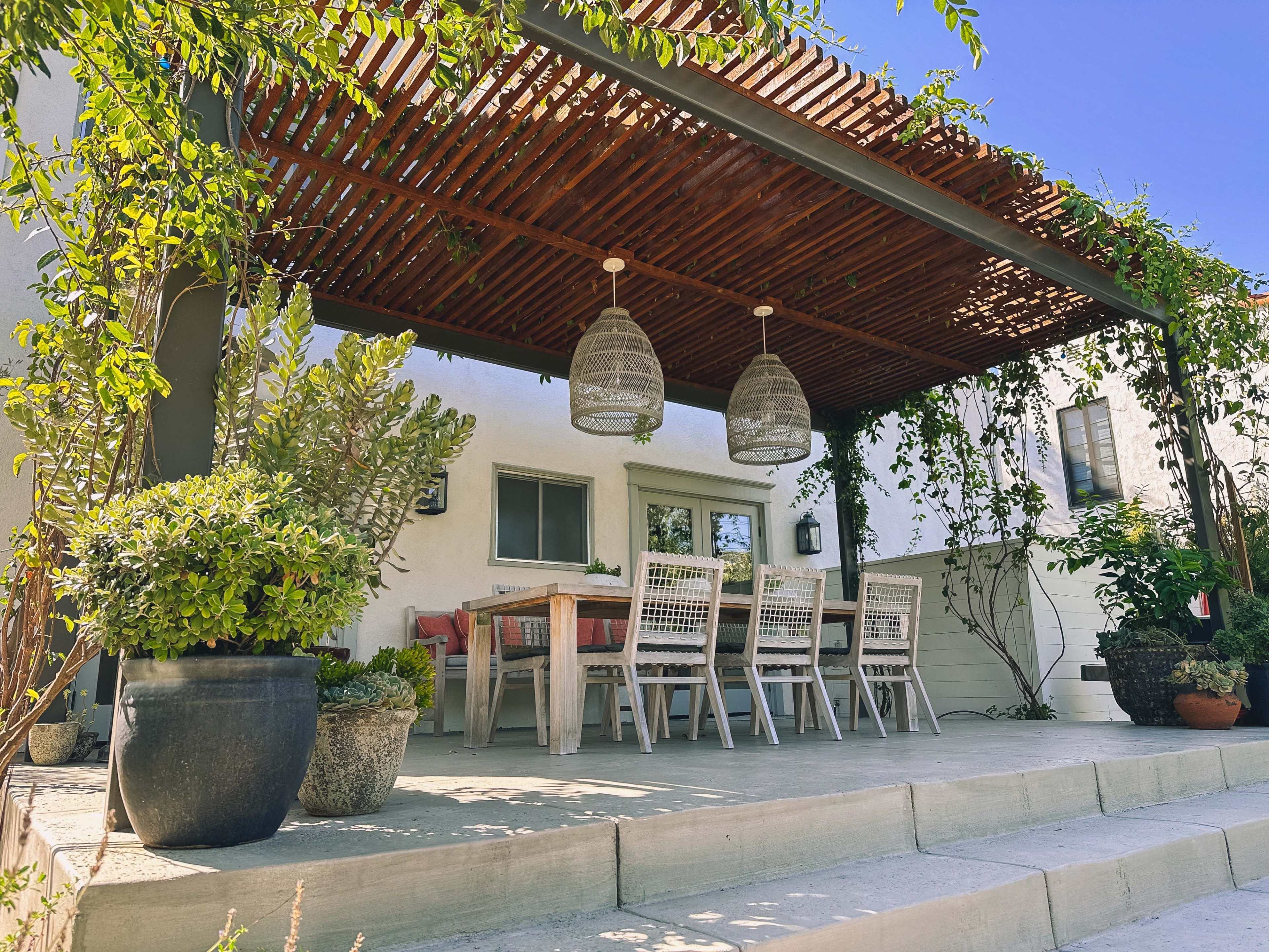 A patio area features a large dining table set beneath a wooden pergola surrounded by potted plants.