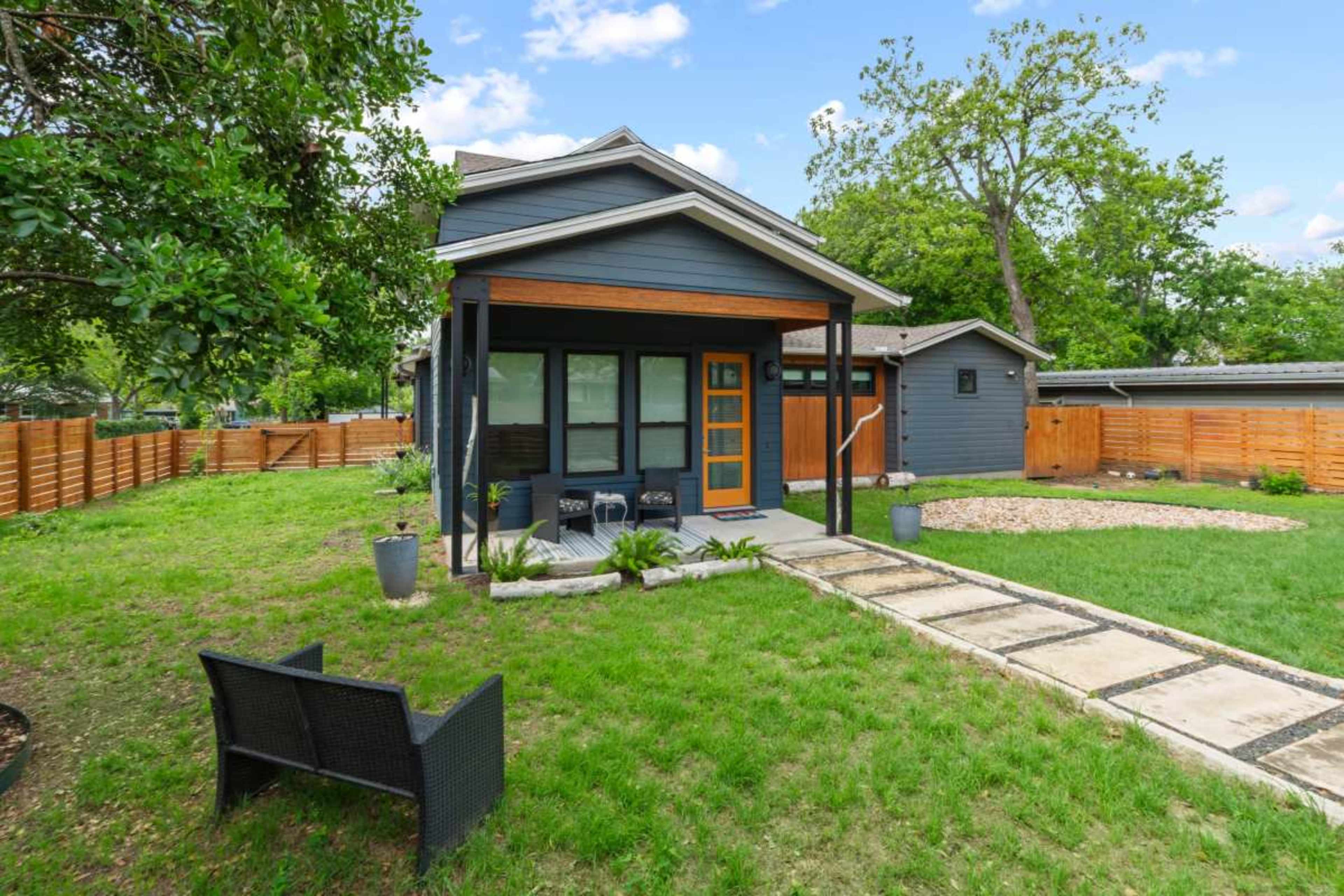 The image shows a backyard featuring a small gray house with wooden accents, a stone pathway, and a neatly landscaped lawn with a seating area.