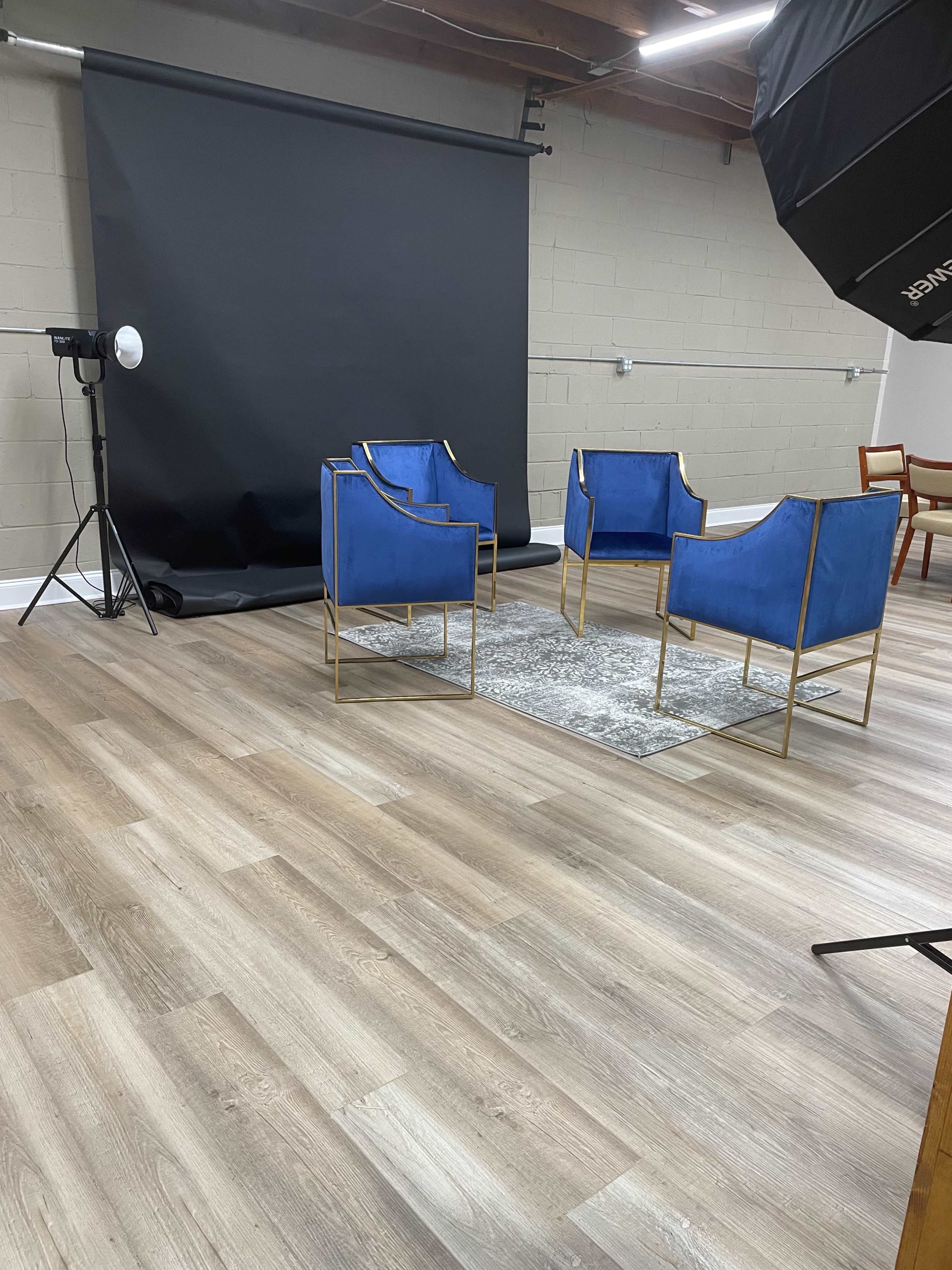 A photography studio featuring four blue chairs arranged on a rug in front of a black backdrop, with a light setup in the corner.