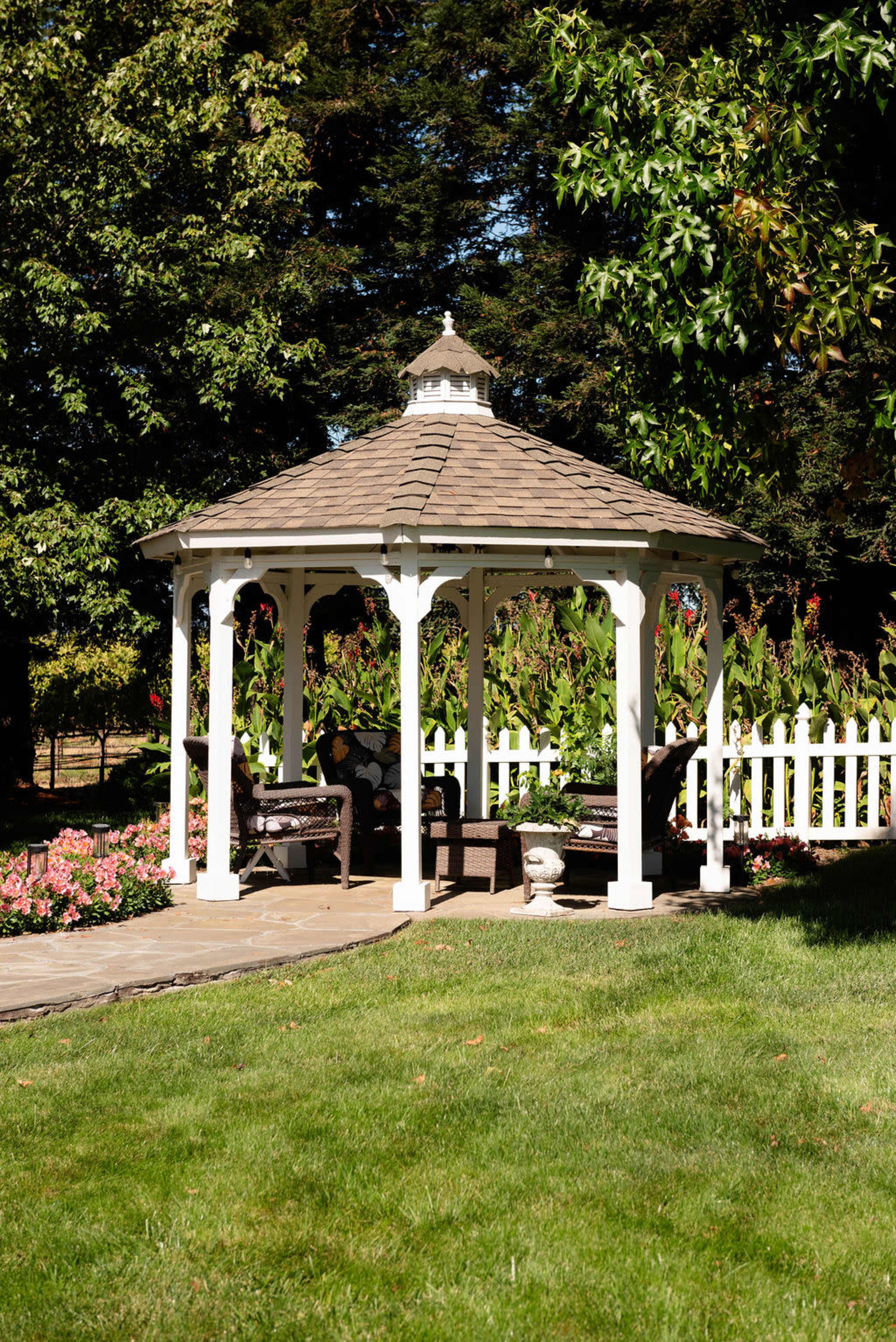 A white gazebo with a shingled roof is surrounded by greenery and flowers in a landscaped garden.