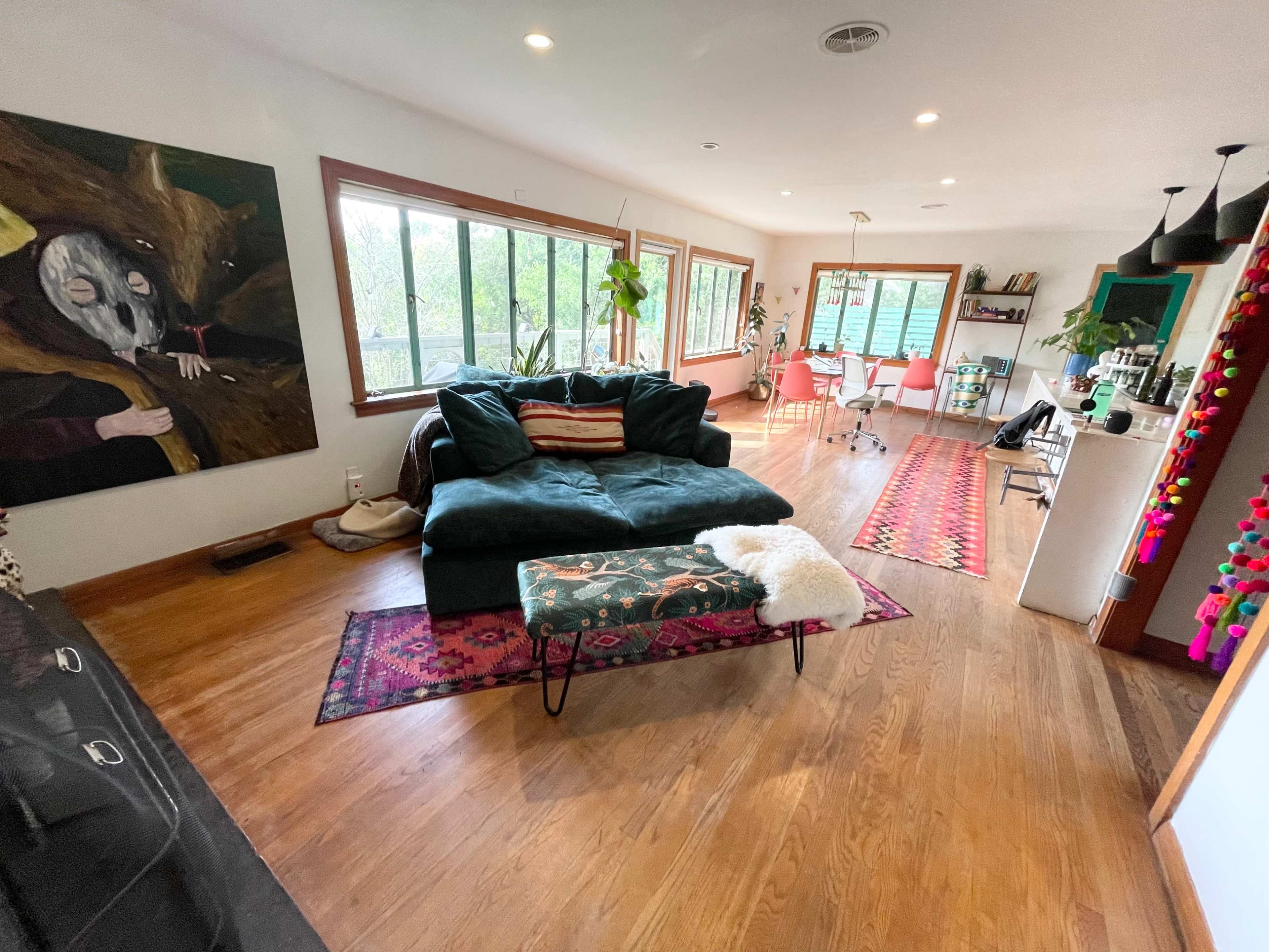 A modern living room with a large dark green sofa, a small bench, and a dining area featuring pink chairs and a large window.