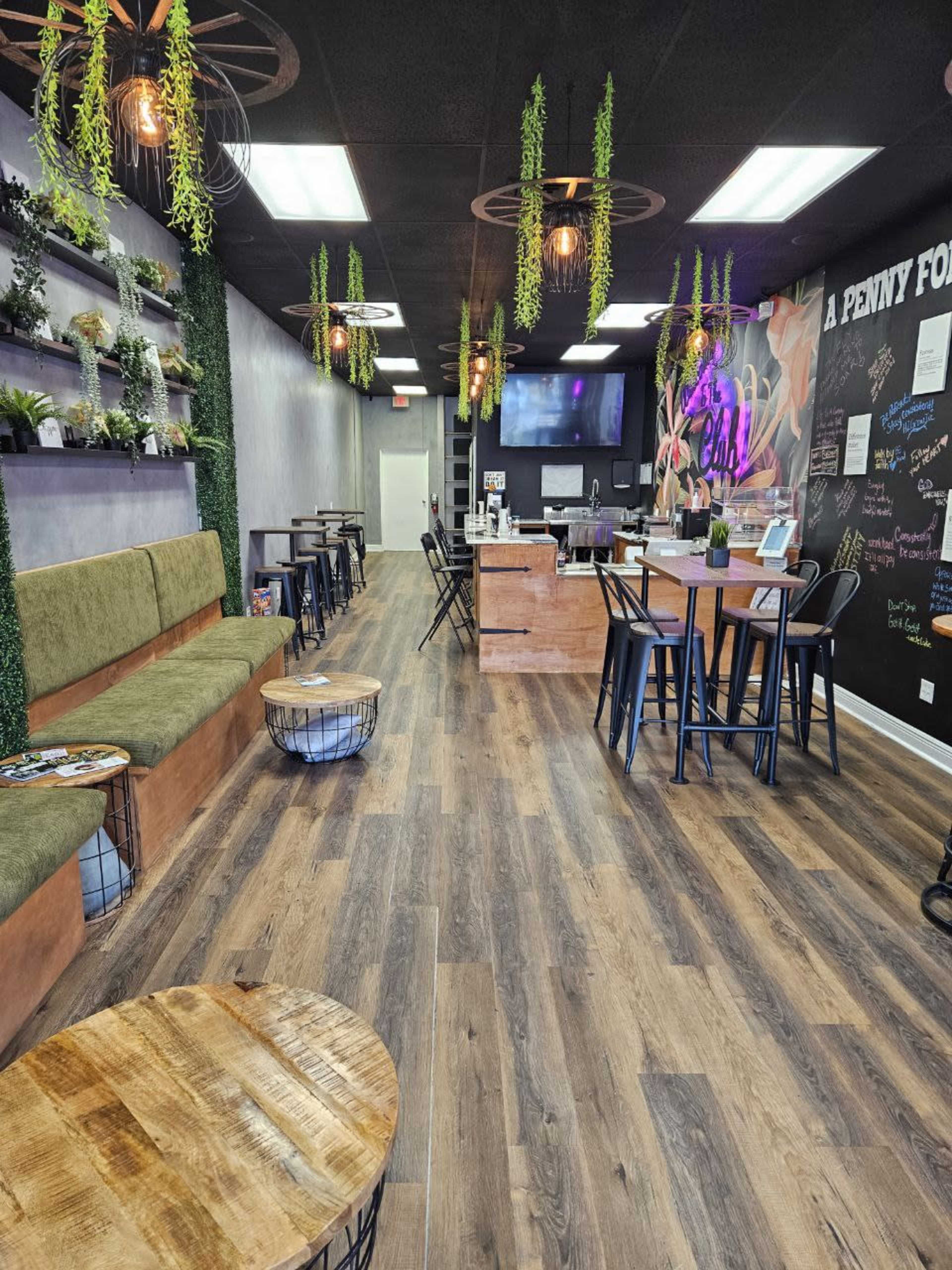 A modern café interior featuring wooden flooring, seating along one side with green cushions, and tables with metal chairs arranged in a spacious layout.