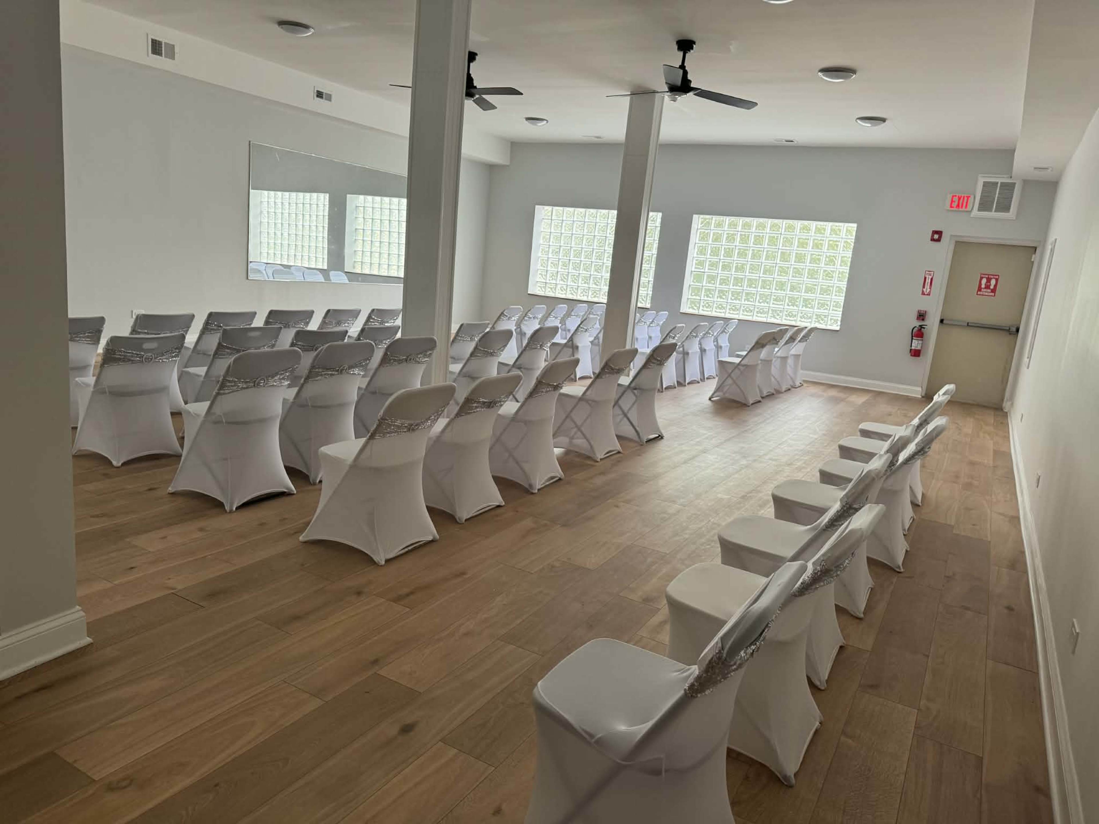 The image shows a modern event space with rows of white chairs, adorned with silver sashes, arranged neatly on a wooden floor.