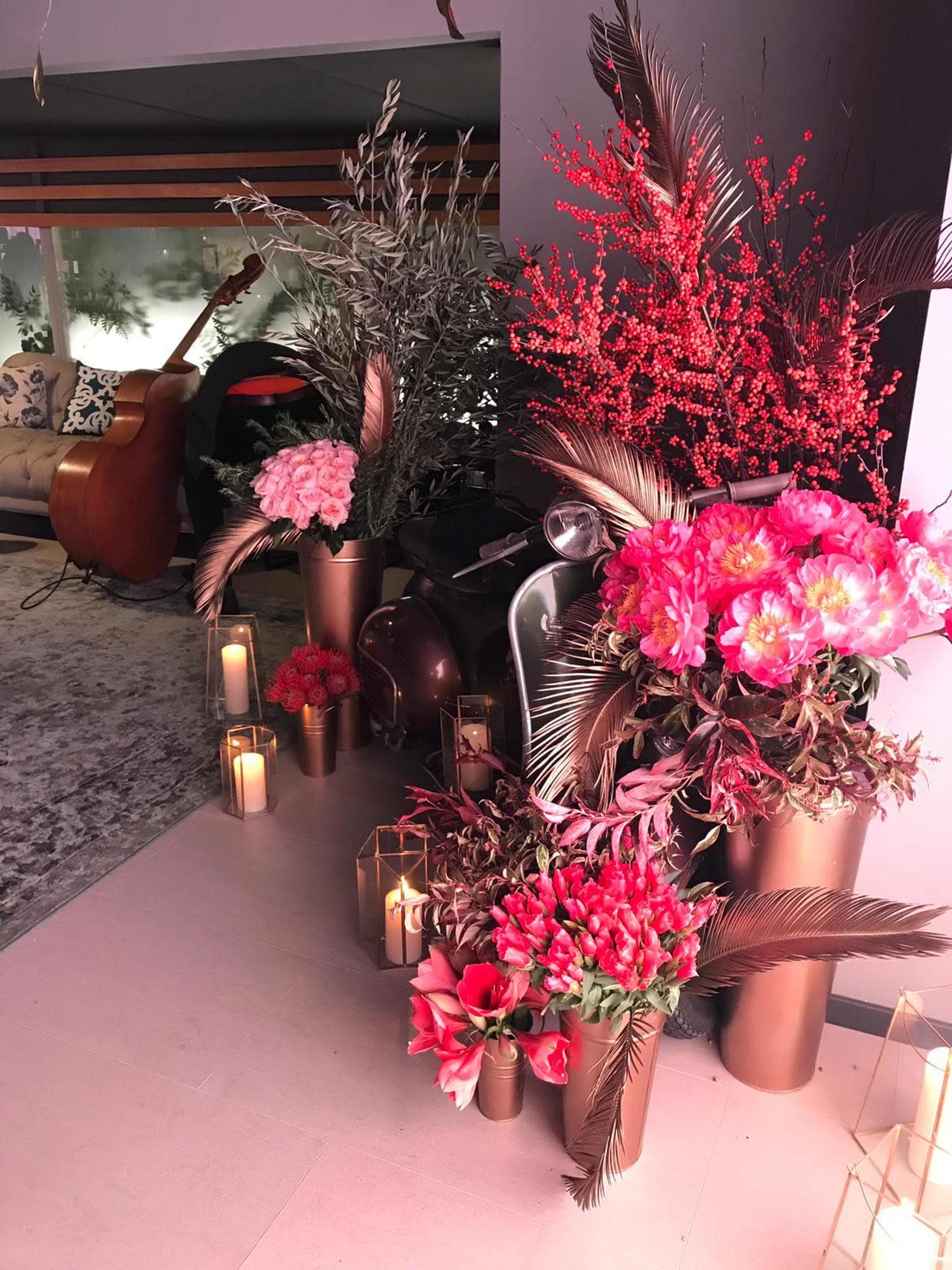 The scene features a collection of vibrant flowers arranged in metallic vases, accompanied by candles, set against a background with a guitar and a sofa.