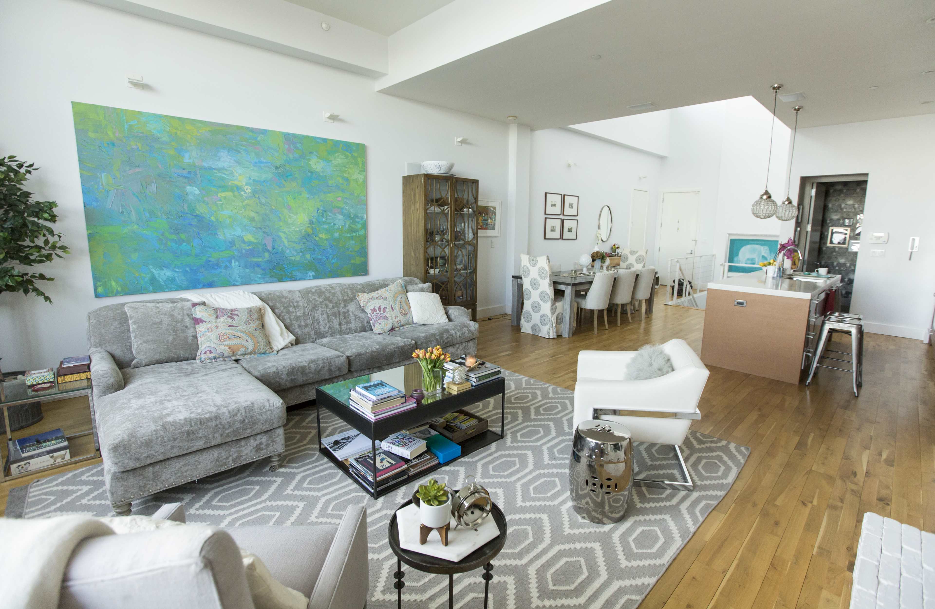 The image shows a modern living room with a gray sofa, a coffee table filled with books, an art piece on the wall, and a dining area in the background with a table and chairs.