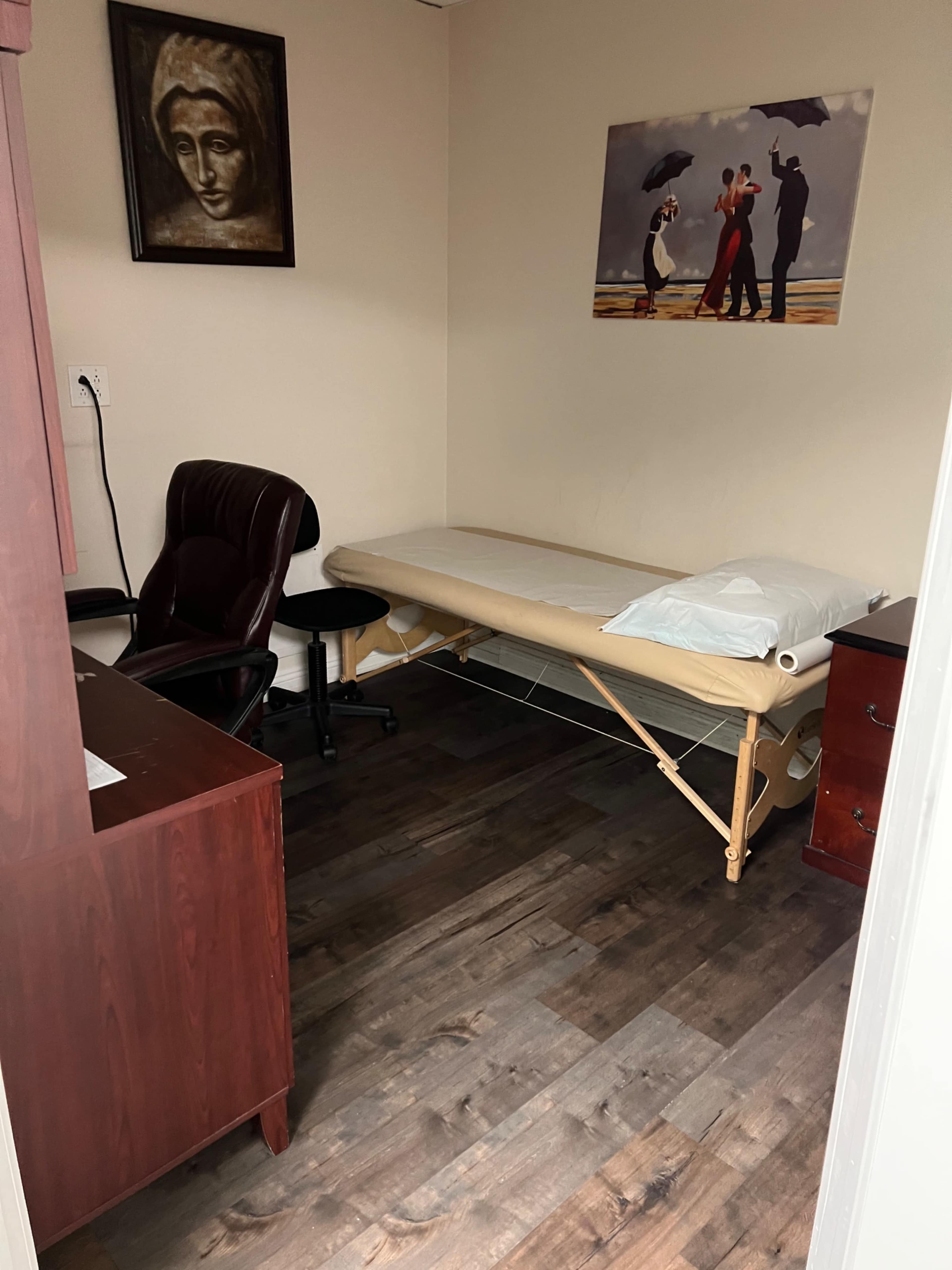 A small treatment room with a wooden floor, a massage table, an office chair, and a desk, along with two framed artworks on the walls.