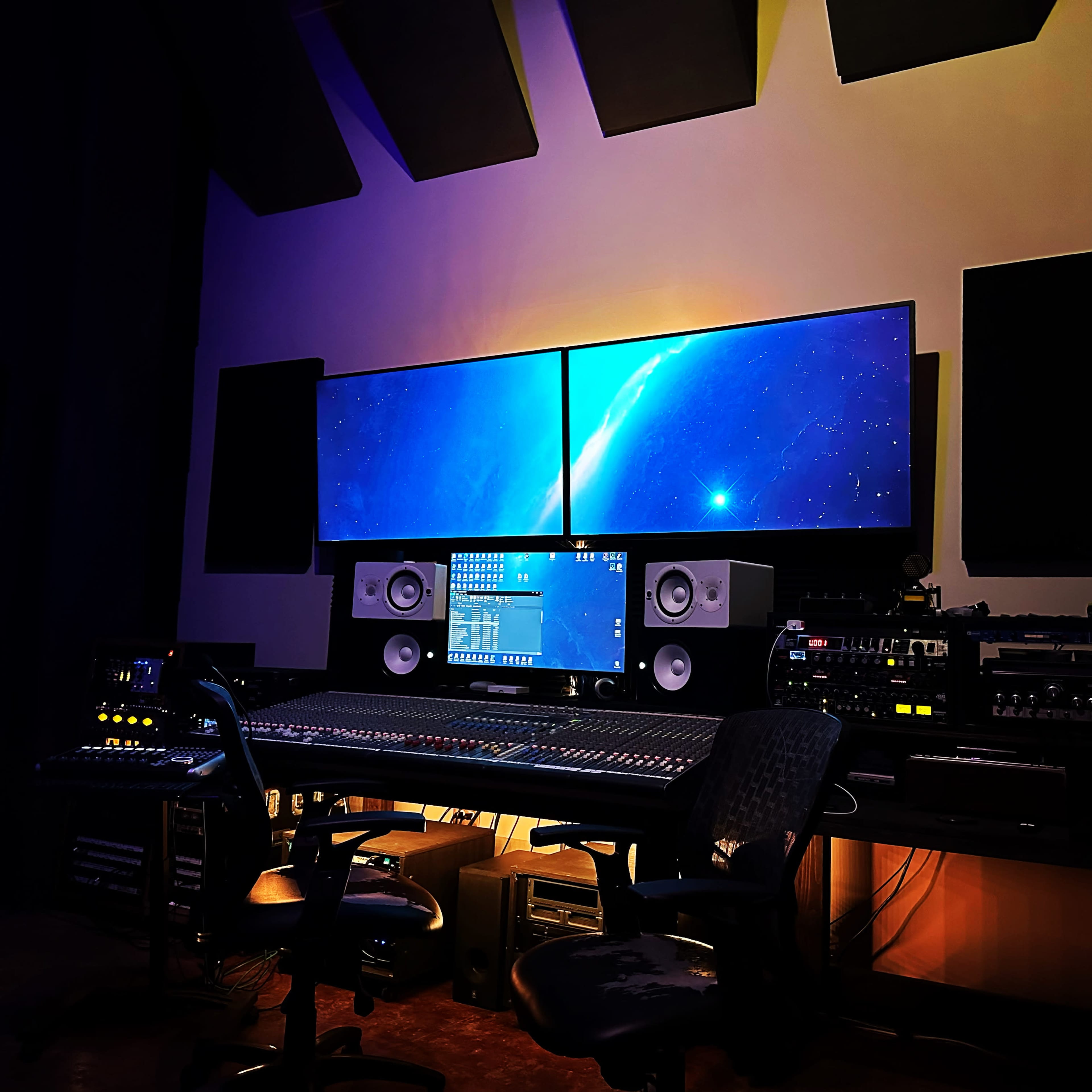 The image shows a music studio with a large mixing console, studio monitors, and two large screens displaying a digital interface.