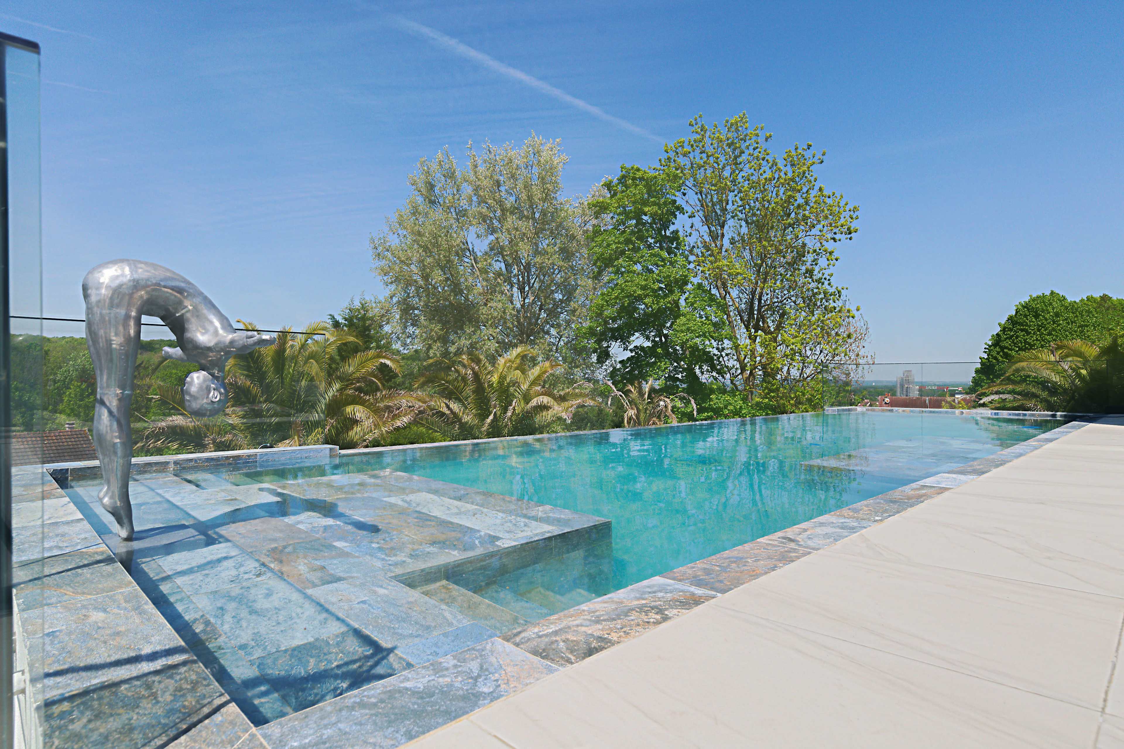 The image shows a modern infinity pool surrounded by greenery and a sculpture of a diver at one end.
