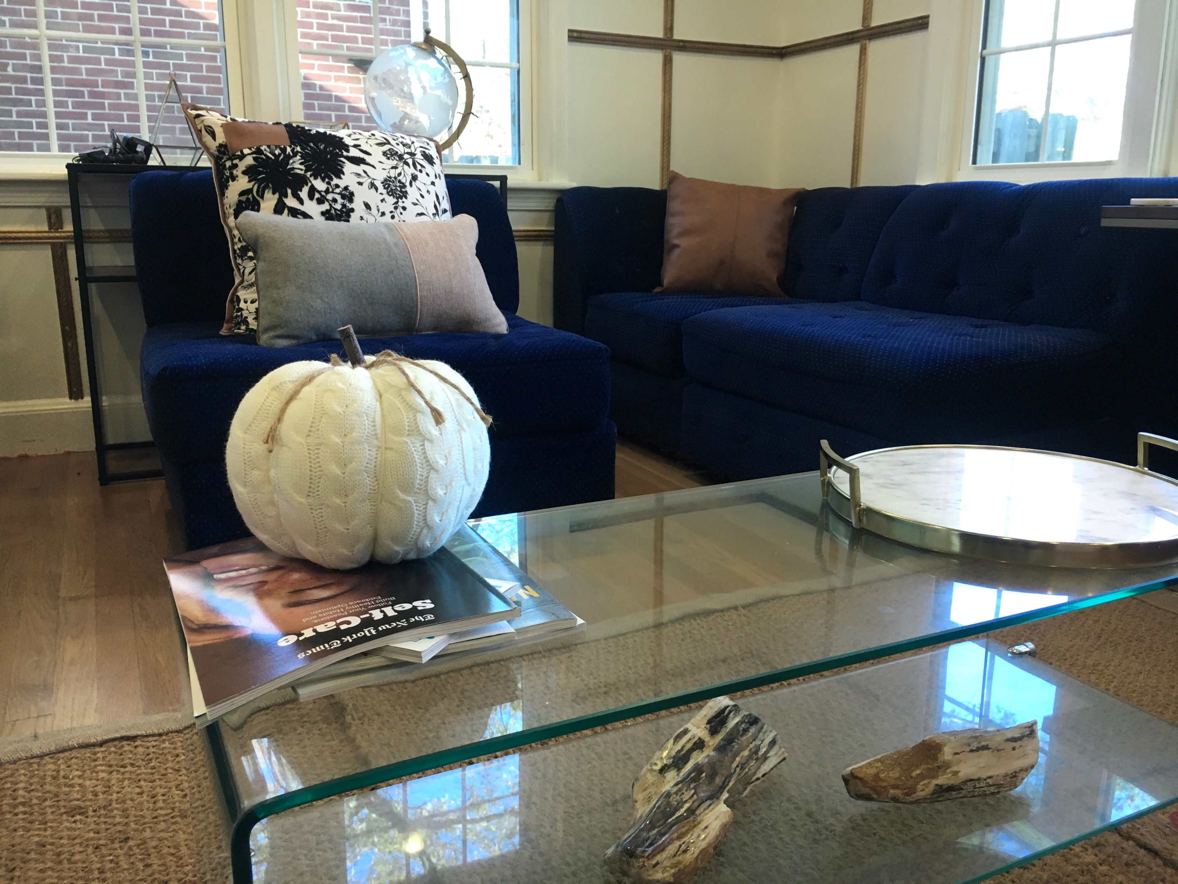 A living room with a blue sectional couch, a glass coffee table with a decorative pumpkin, and large windows providing natural light.
