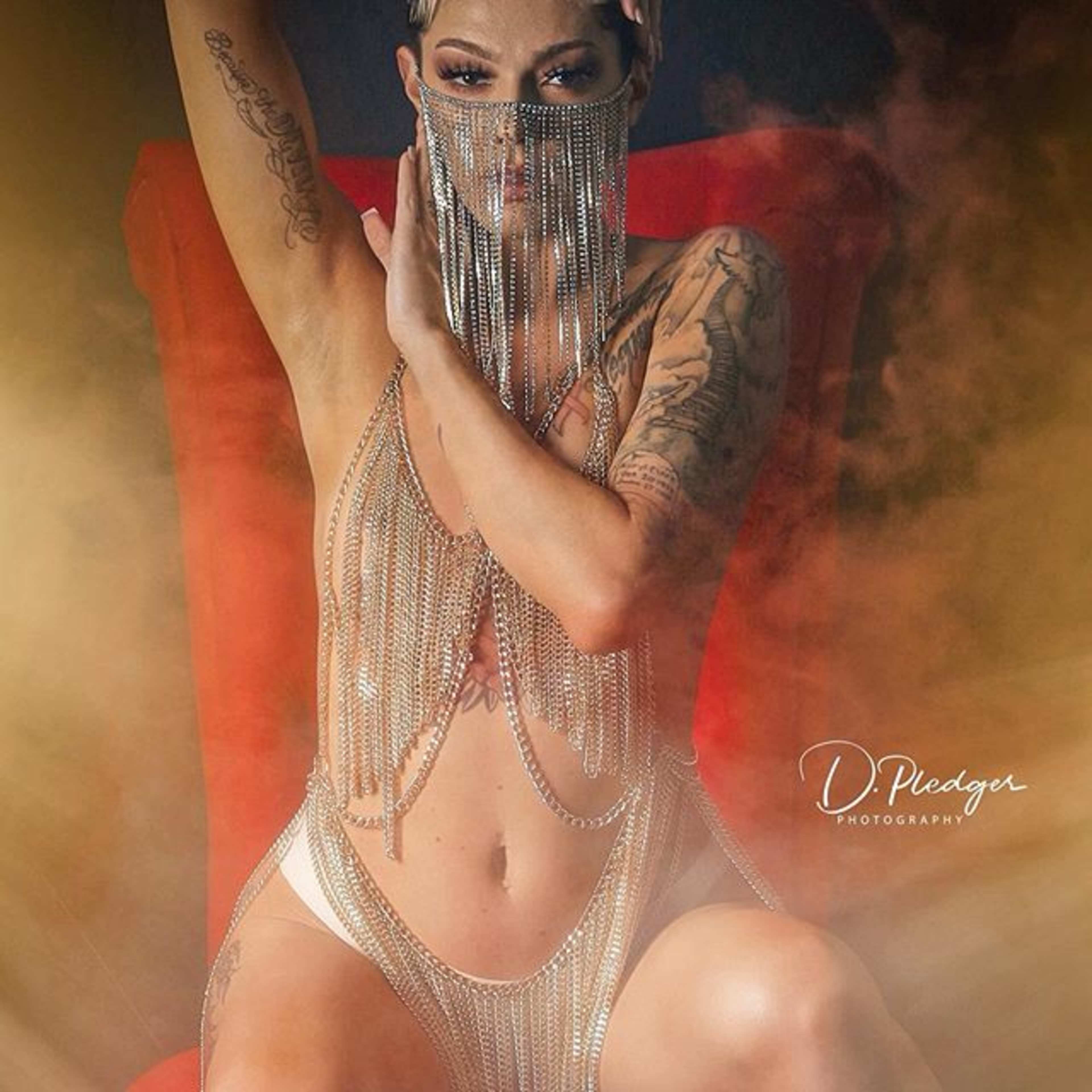 A woman with tattoos poses in a chain-link outfit while seated on a red chair, surrounded by soft smoke.