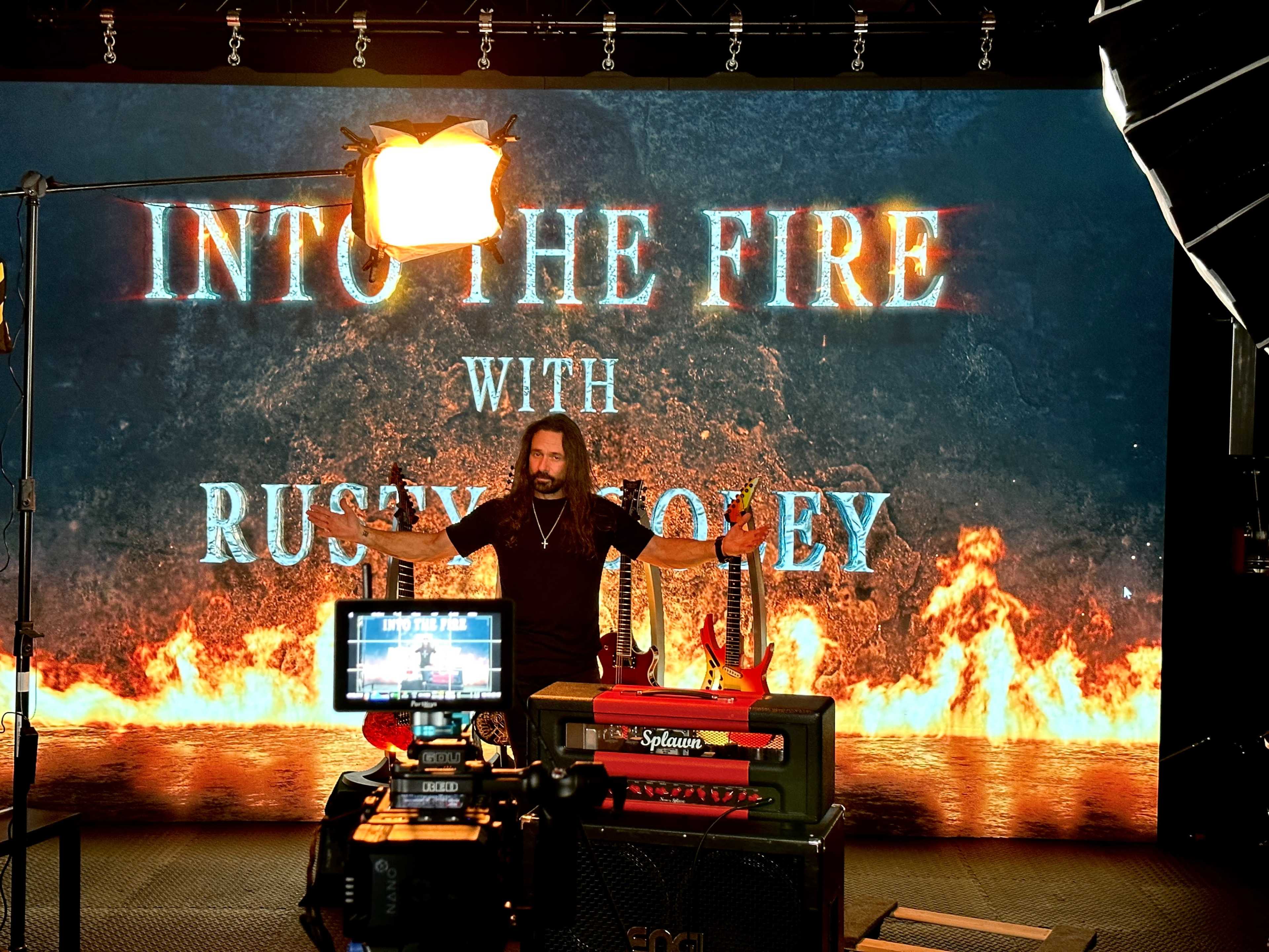 A man stands in front of a large screen displaying the title "Into the Fire with Rusty Mosley," surrounded by musical equipment and a camera.