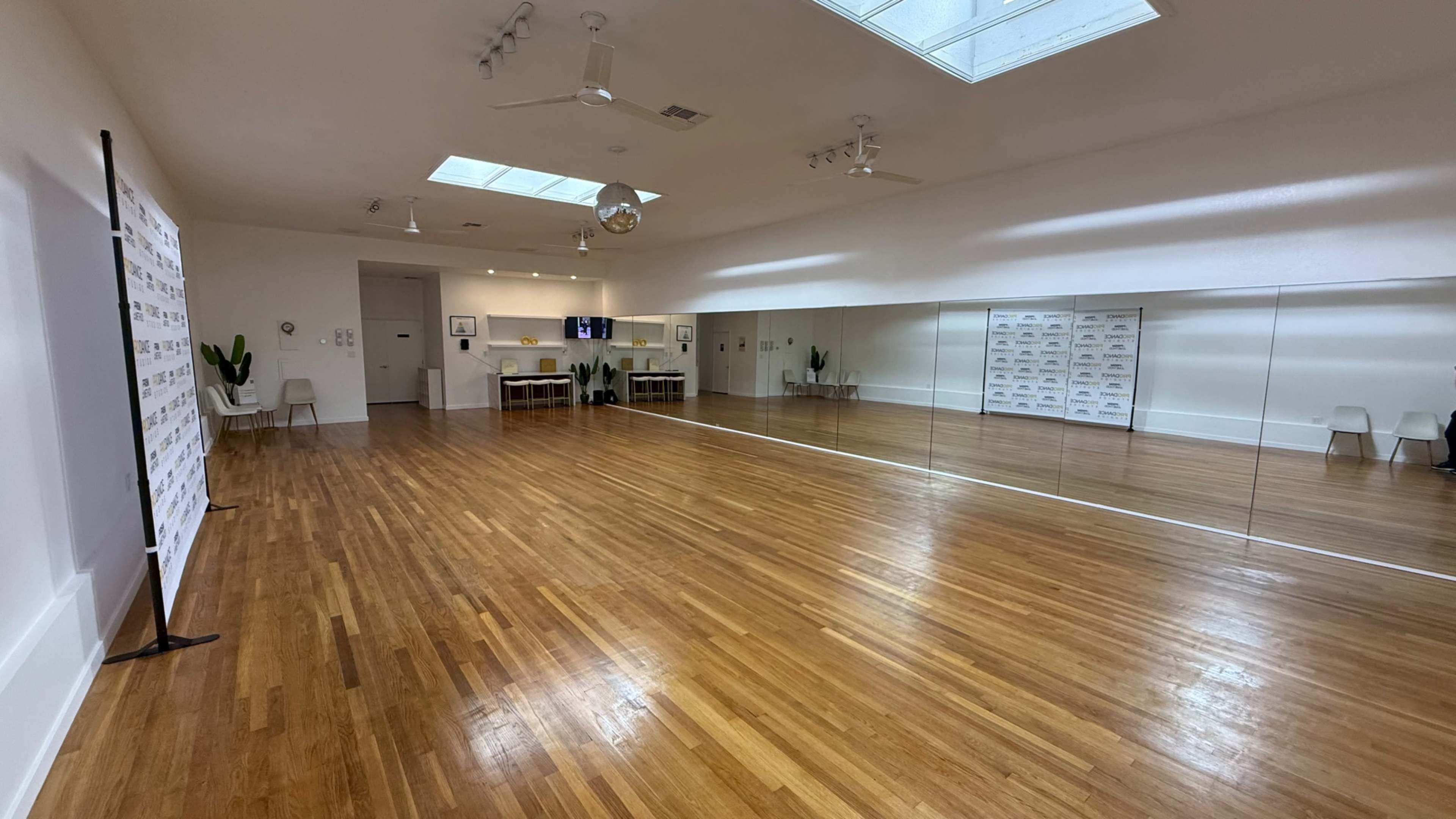 The image shows a spacious, well-lit dance studio with polished wooden floors, large mirrors on one wall, and a minimalist design.