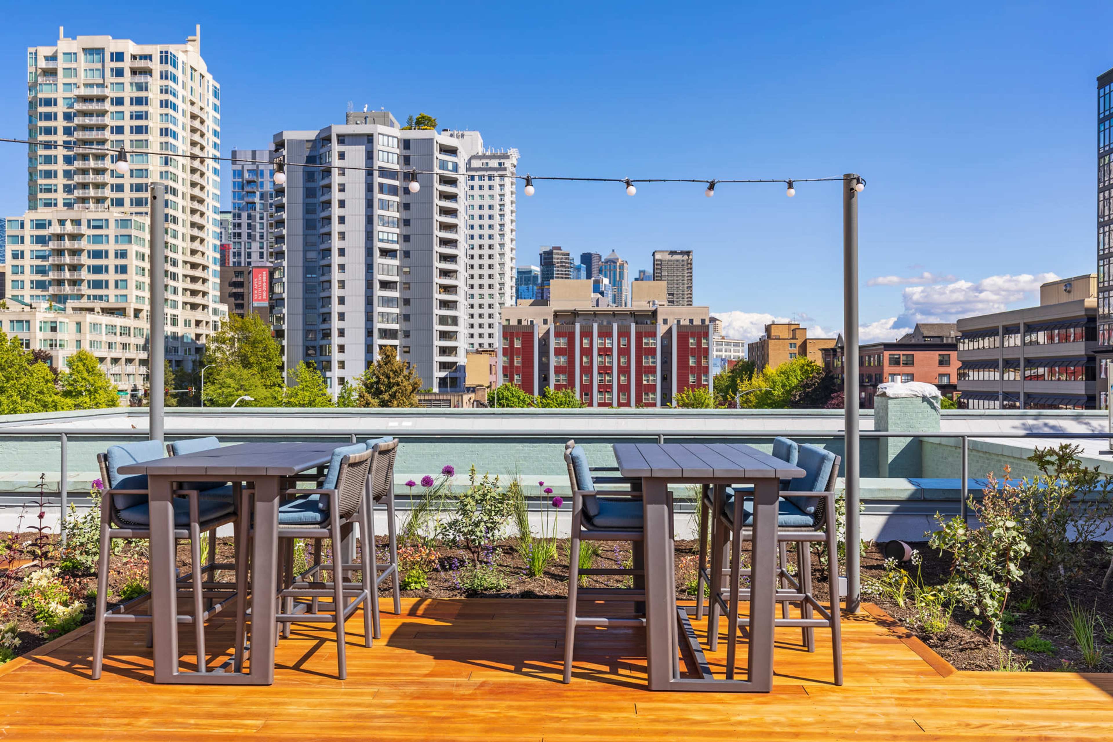 A rooftop terrace with grey furniture and string lights, overlooking a cityscape of tall buildings and clear blue skies.