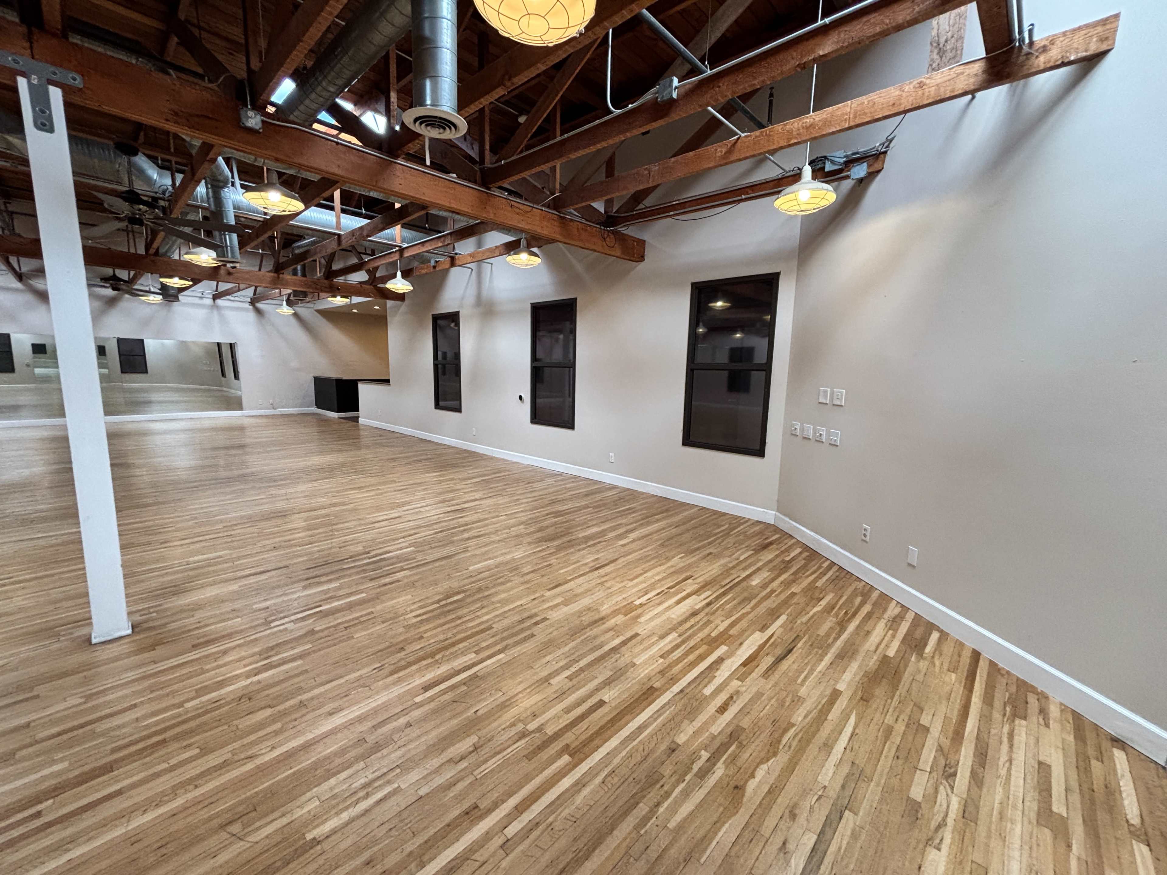 The image shows a spacious, empty room with wooden flooring, exposed beams, and several windows.