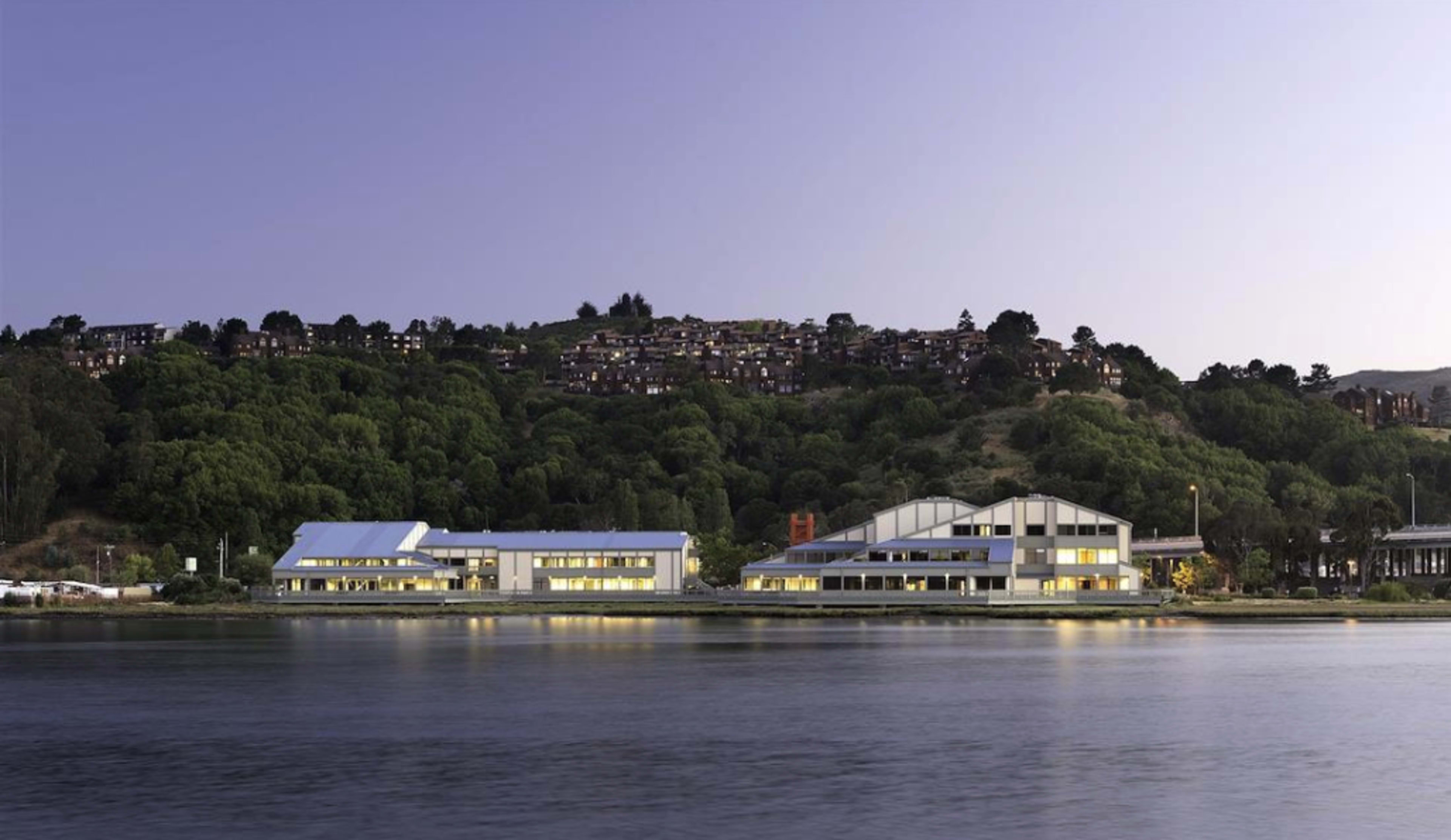 A modern building complex is situated along the water, with a hillside of homes in the background under a twilight sky.