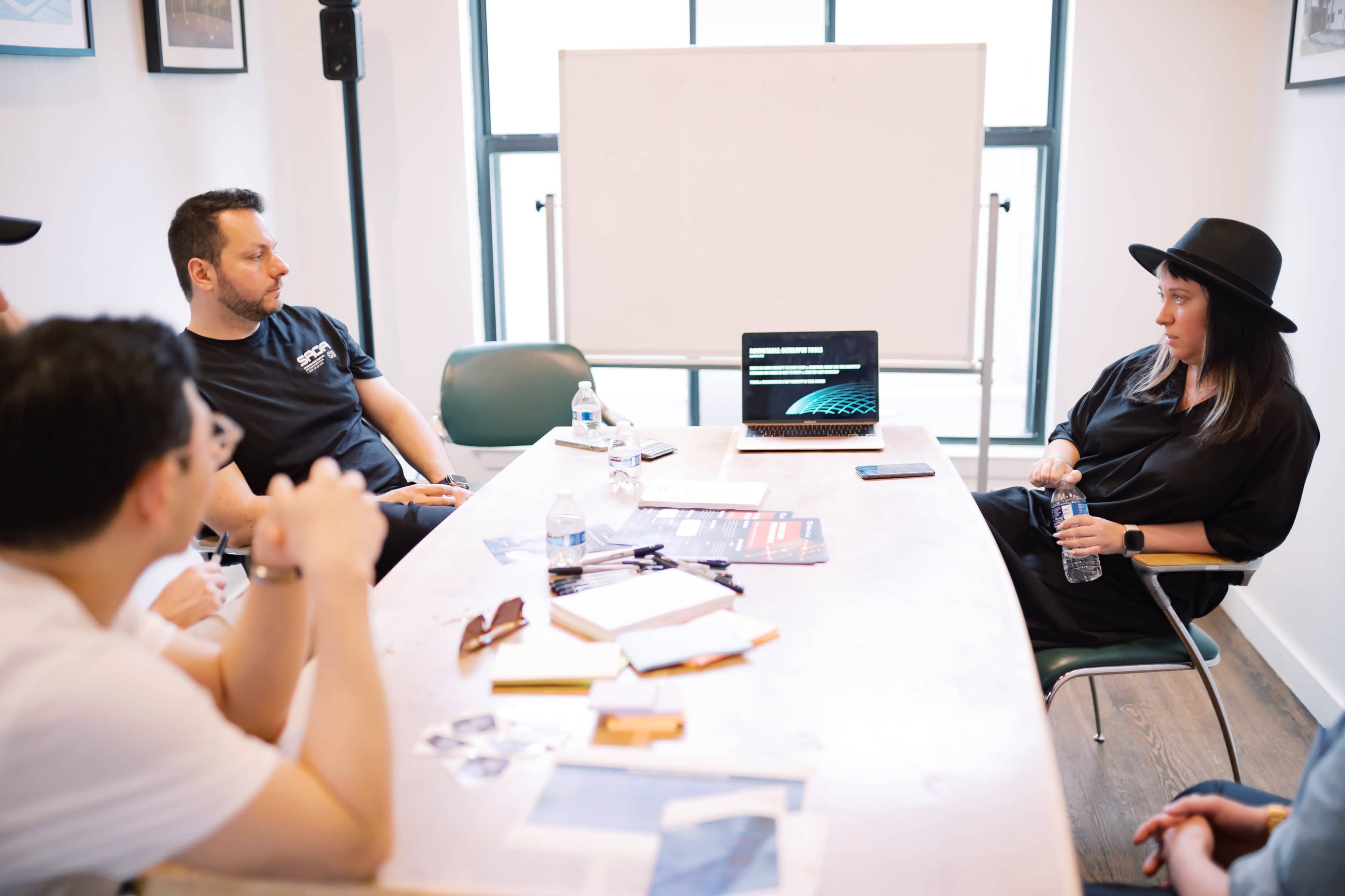 A group of people is seated around a wooden table in a meeting room, with a laptop displaying a presentation and notes visible on the table.