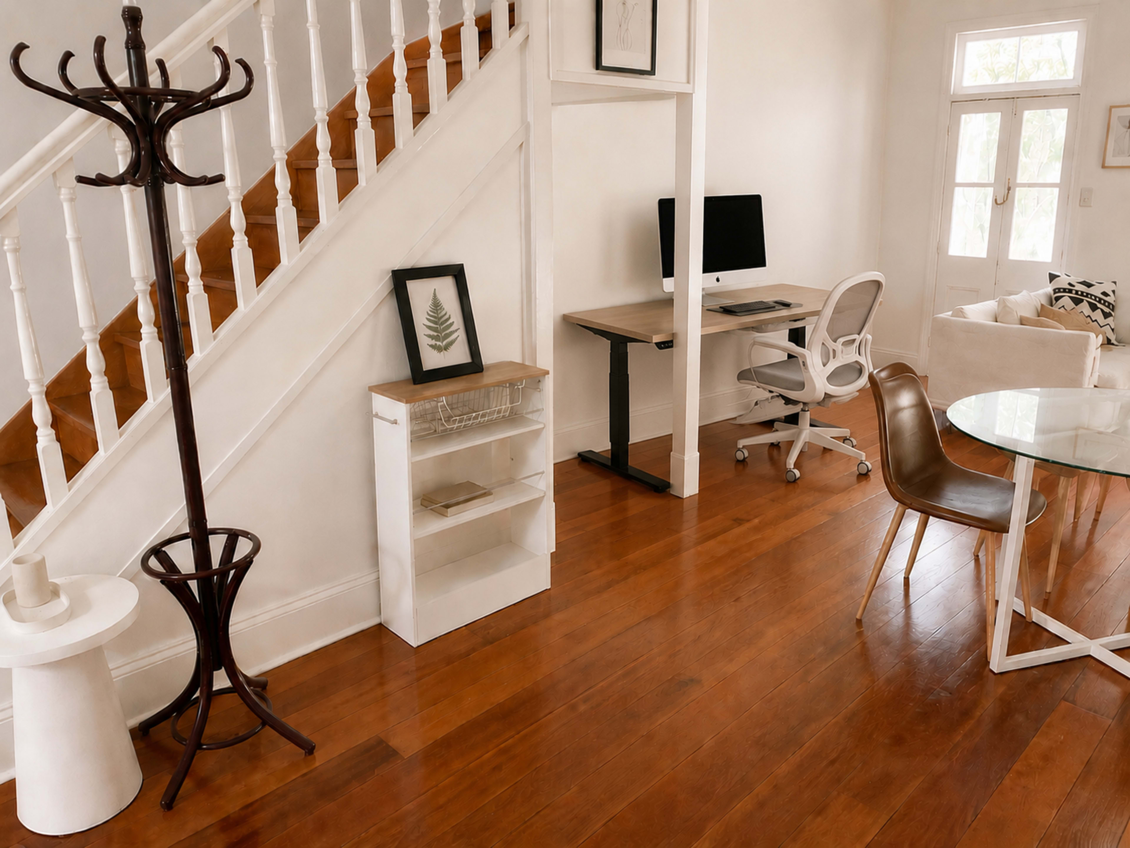 The image shows a modern interior space featuring a staircase, a coat rack, a small shelf, a desk with a computer, and a dining table set.
