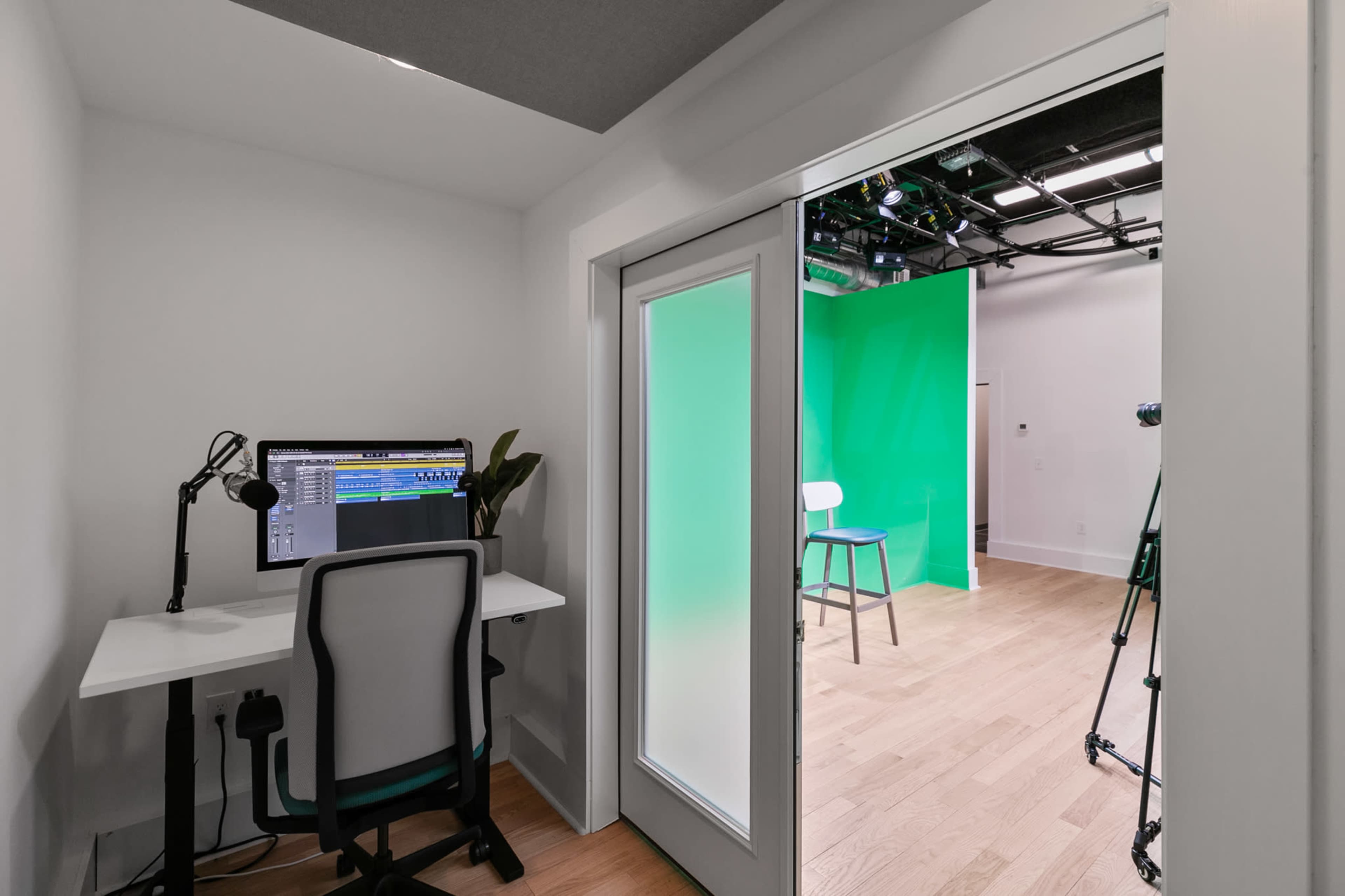 A small room features a desk with a computer and microphone, leading into a studio space with green backdrop and lighting equipment.